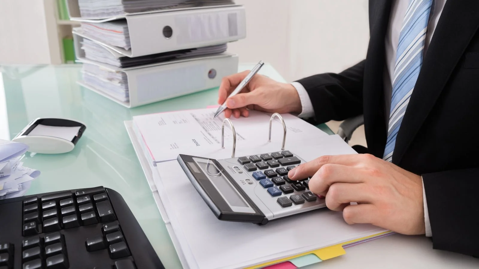 Businessperson in a suit using a calculator and writing on documents at a cluttered office desk.