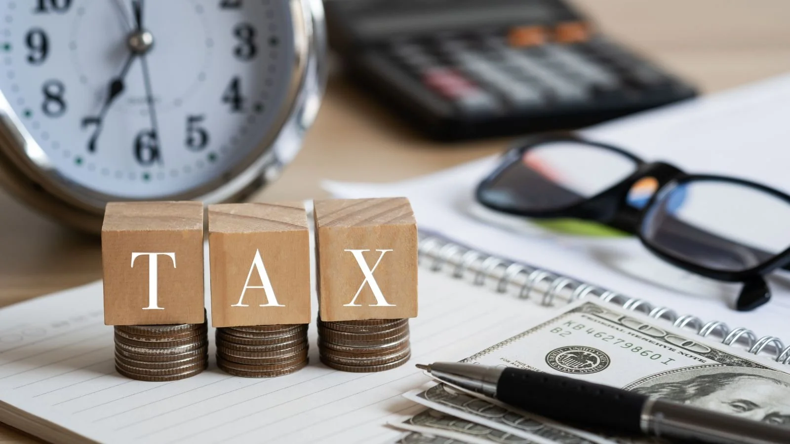 Wooden blocks spelling 'TAX' placed on stacks of coins on a notebook, with a clock, calculator, glasses, a pen, and dollar bills in the background.