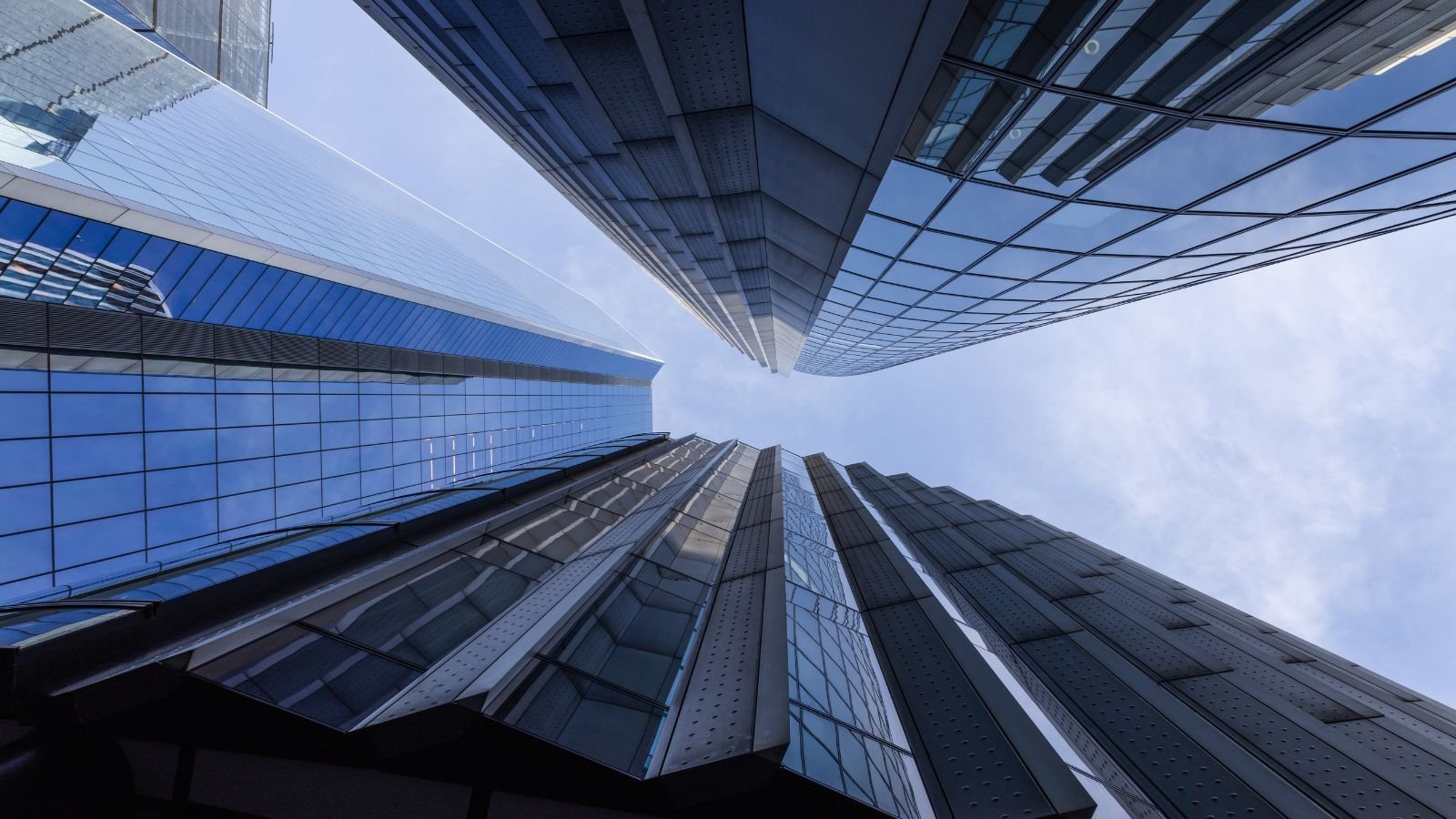 Looking up at tall modern glass skyscrapers from the ground, showing reflections of the sky and adjacent buildings.