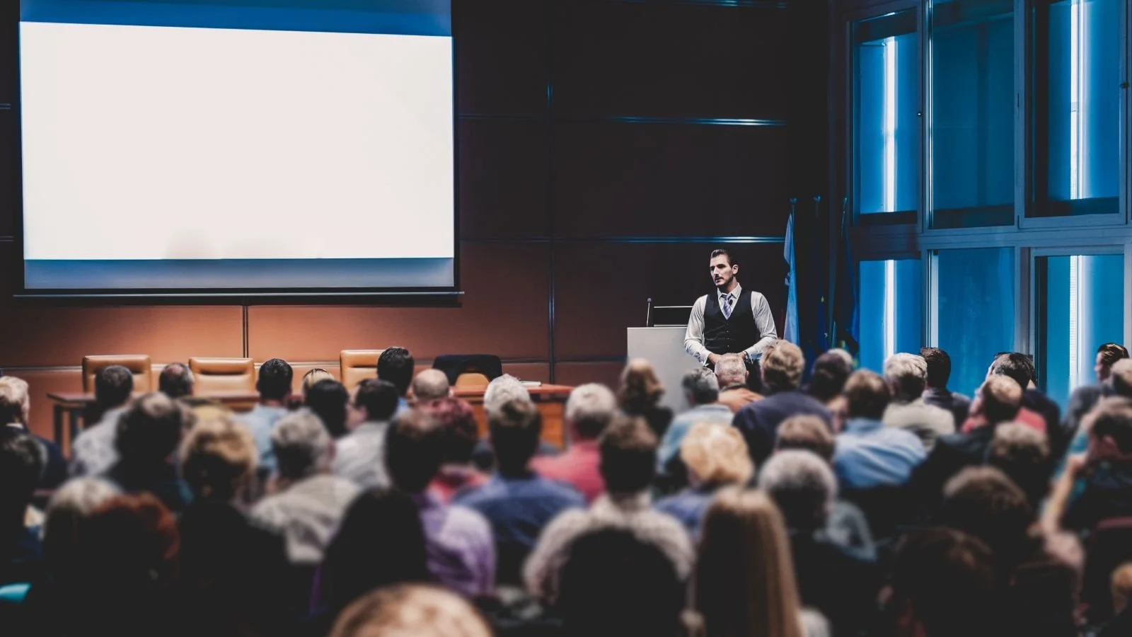 A man in formal attire giving a presentation to an audience in a conference room with a large screen behind him.