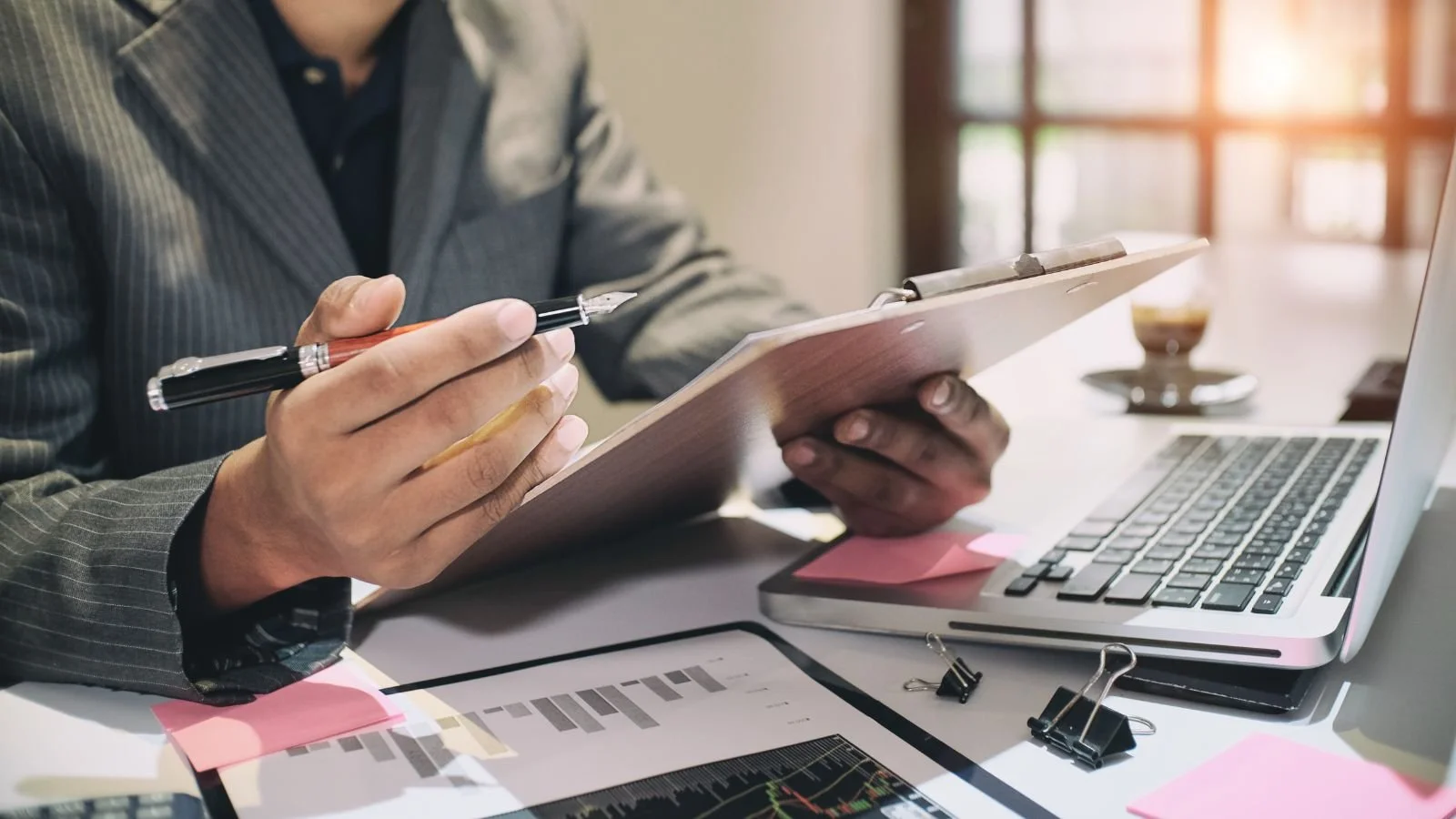 A person in a business suit holds a clipboard and pen, working at a desk with a laptop, financial charts, and office supplies, in a bright office.