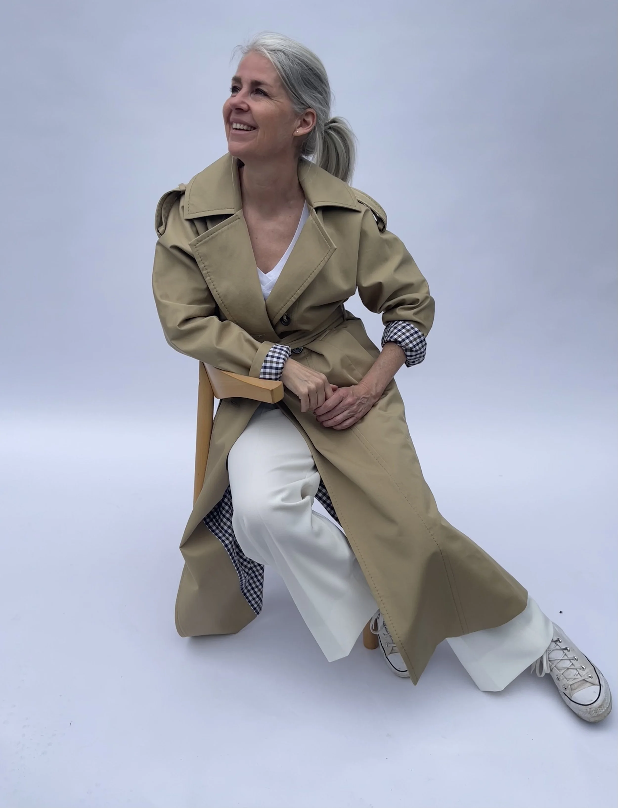An older woman with gray hair tied back in a ponytail, smiling while sitting on a chair against a plain light background. She is wearing a tan trench coat, white pants, plaid shirt cuffs visible, and beige sneakers.