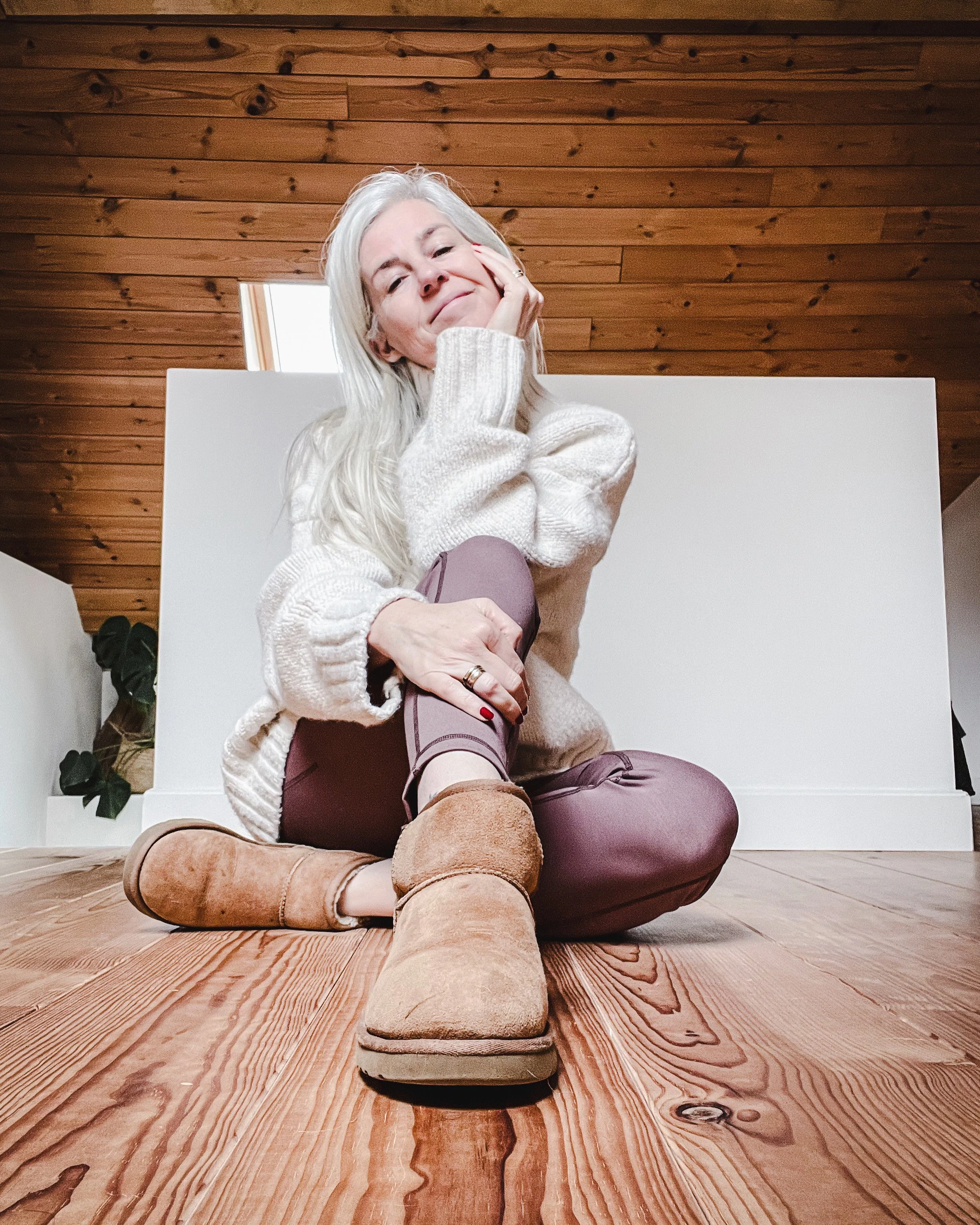A woman with long silver hair sitting on a wooden floor, smiling and touching her face, wearing a beige sweater, mauve pants, and tan boots, in a room with a wooden ceiling and a small green plant in the background.
