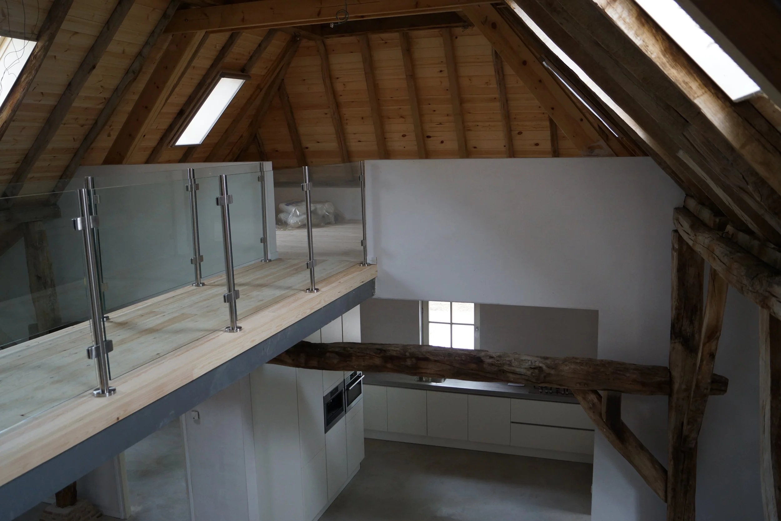 Interior view of a loft with wooden ceiling and beams, glass balcony railing, kitchen with white cabinets and a window.