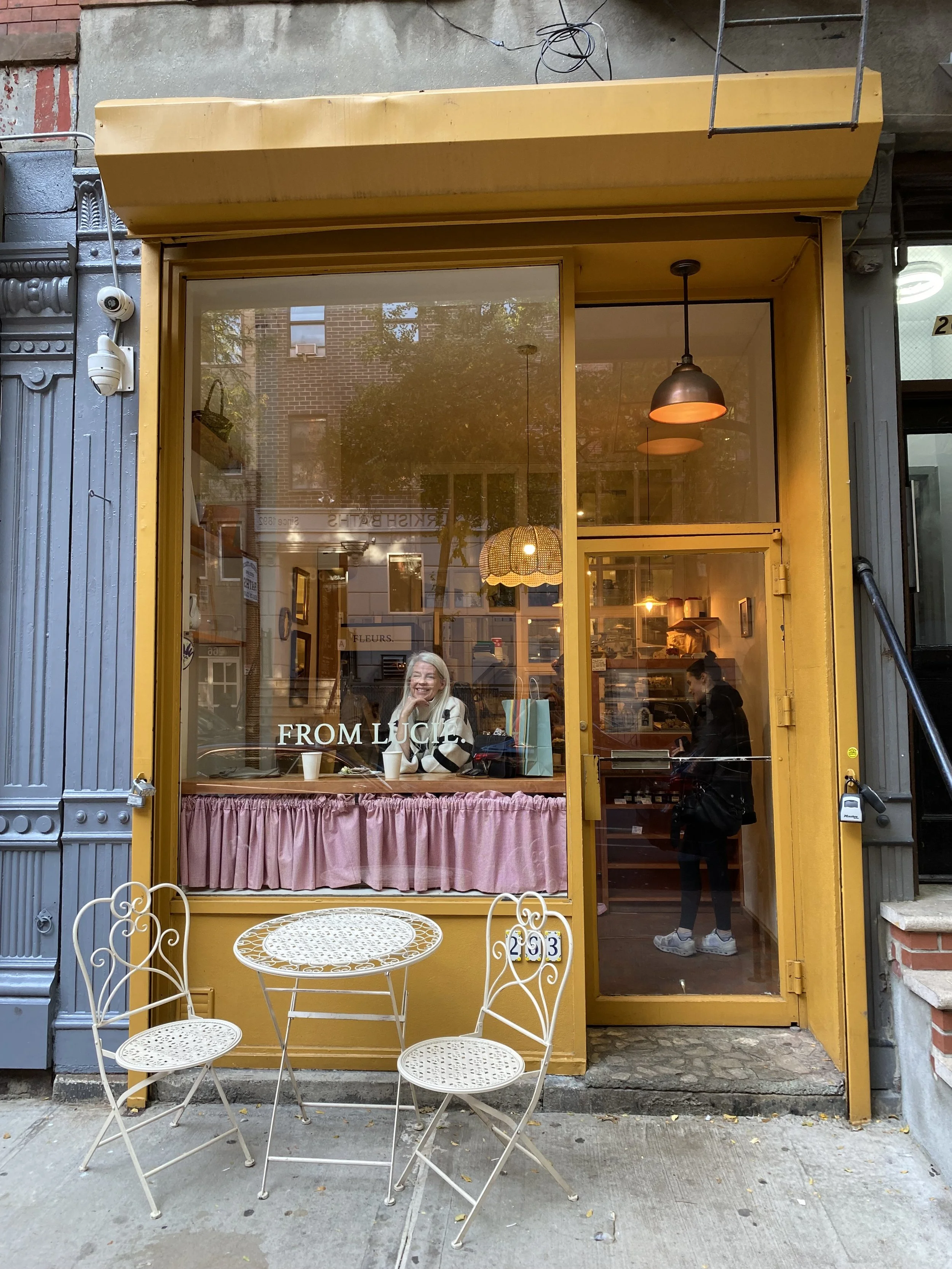 A small cafe with a yellow exterior, a large front window, and a chalkboard sign. Outside, there are two white metal chairs and a small round table on the sidewalk. Inside, a woman is sitting at the window with a drink, smiling, and another person is inside near the counter.