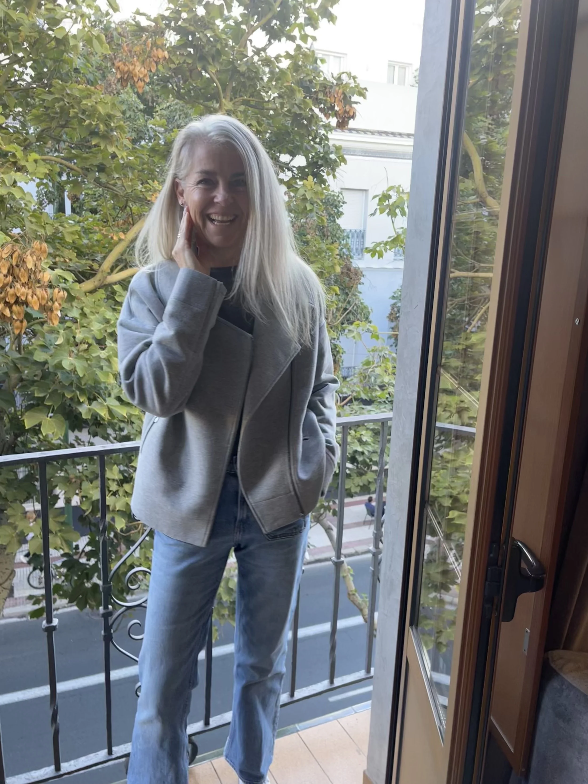 A woman with long blonde hair smiling and standing on a small balcony by a glass door. She is wearing a light gray jacket and blue jeans, with trees and buildings visible in the background.