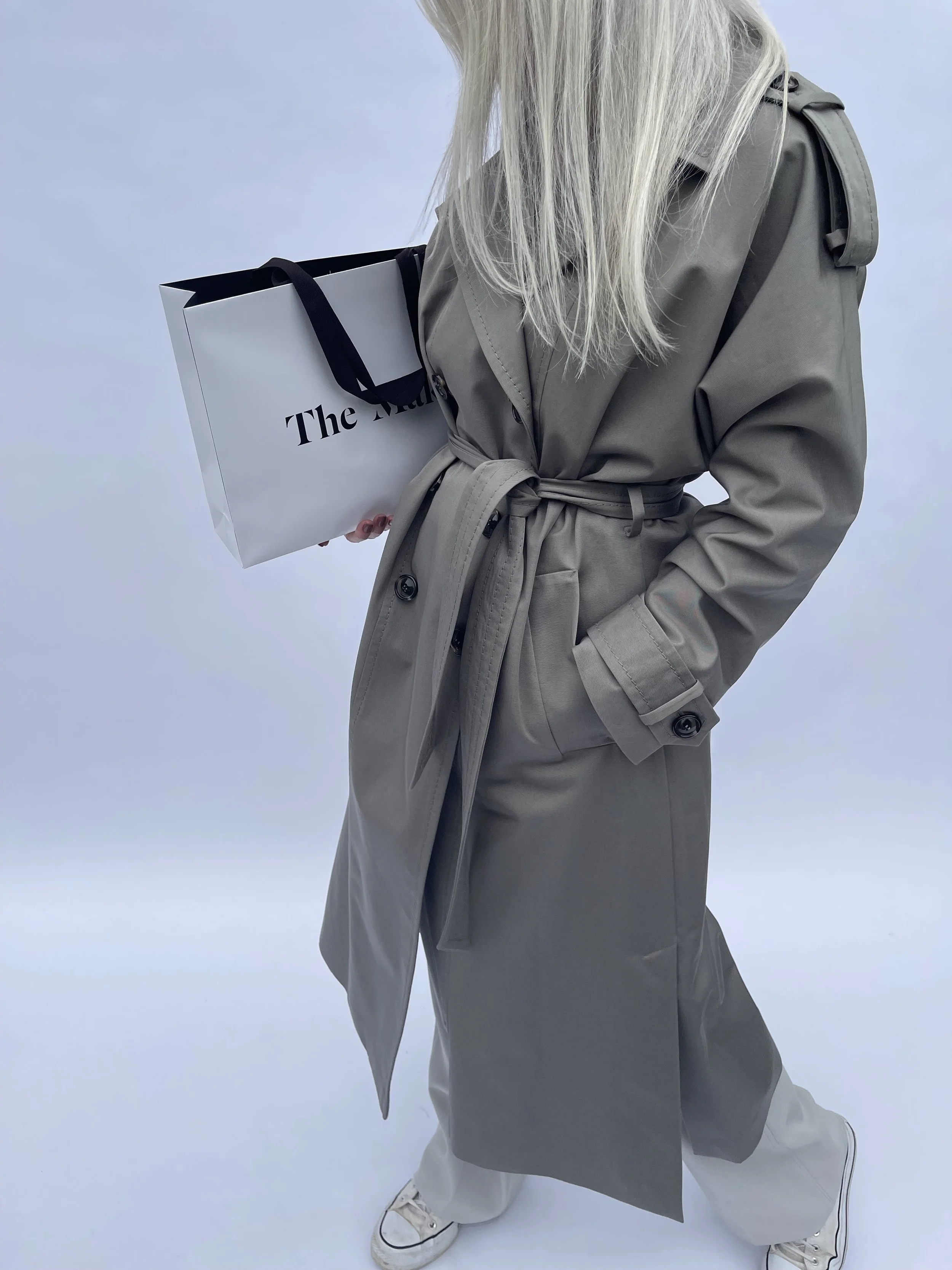 A woman with long blonde hair wearing a beige trench coat, light-colored pants, and white sneakers, holding a white shopping bag with black handles that has partially visible text, standing against a plain light background.