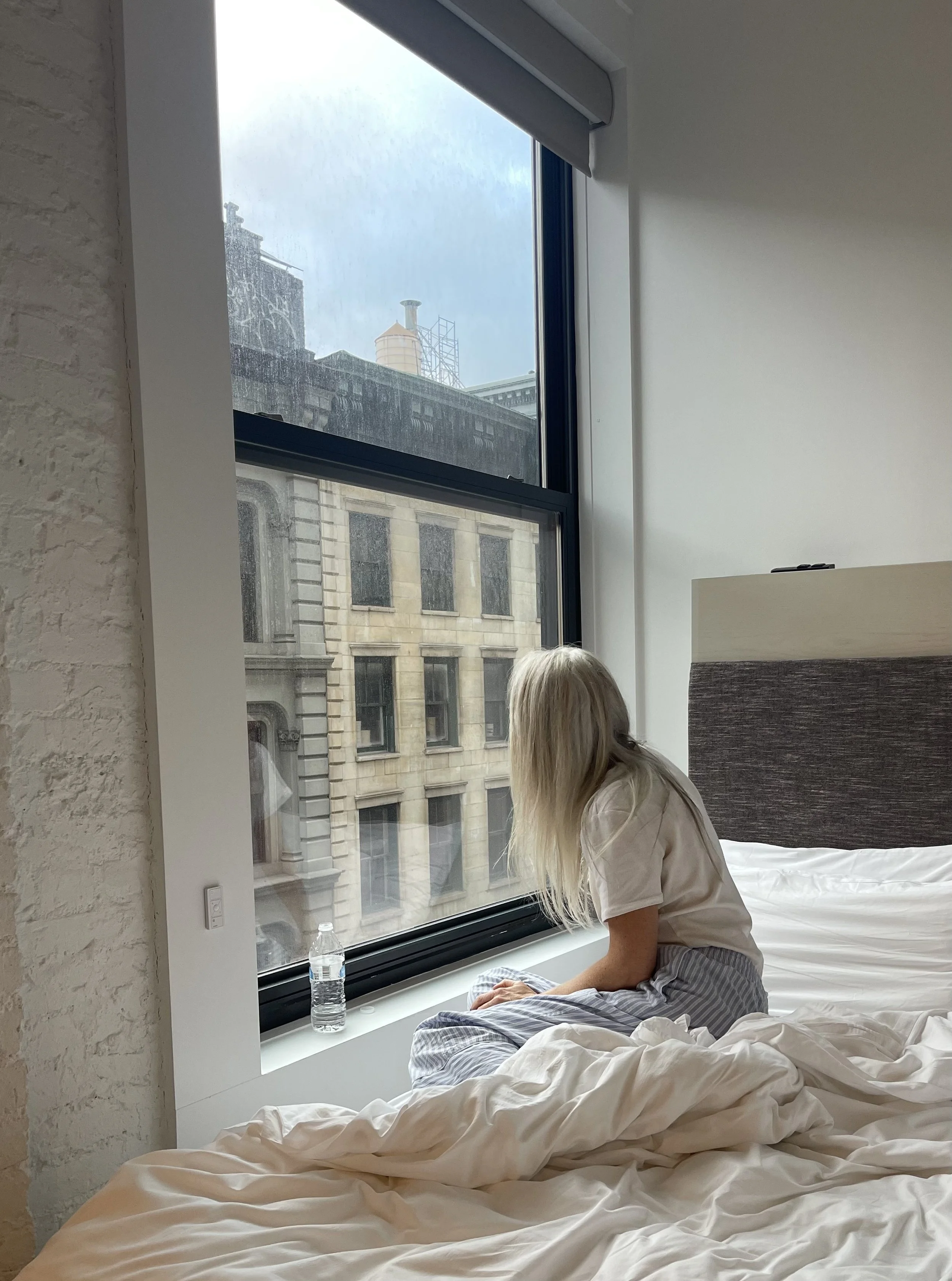 A woman with long blonde hair sitting on a bed near a large window, looking outside at the city buildings and sky, with a water bottle placed on the window sill, in a modern, minimalistic bedroom.