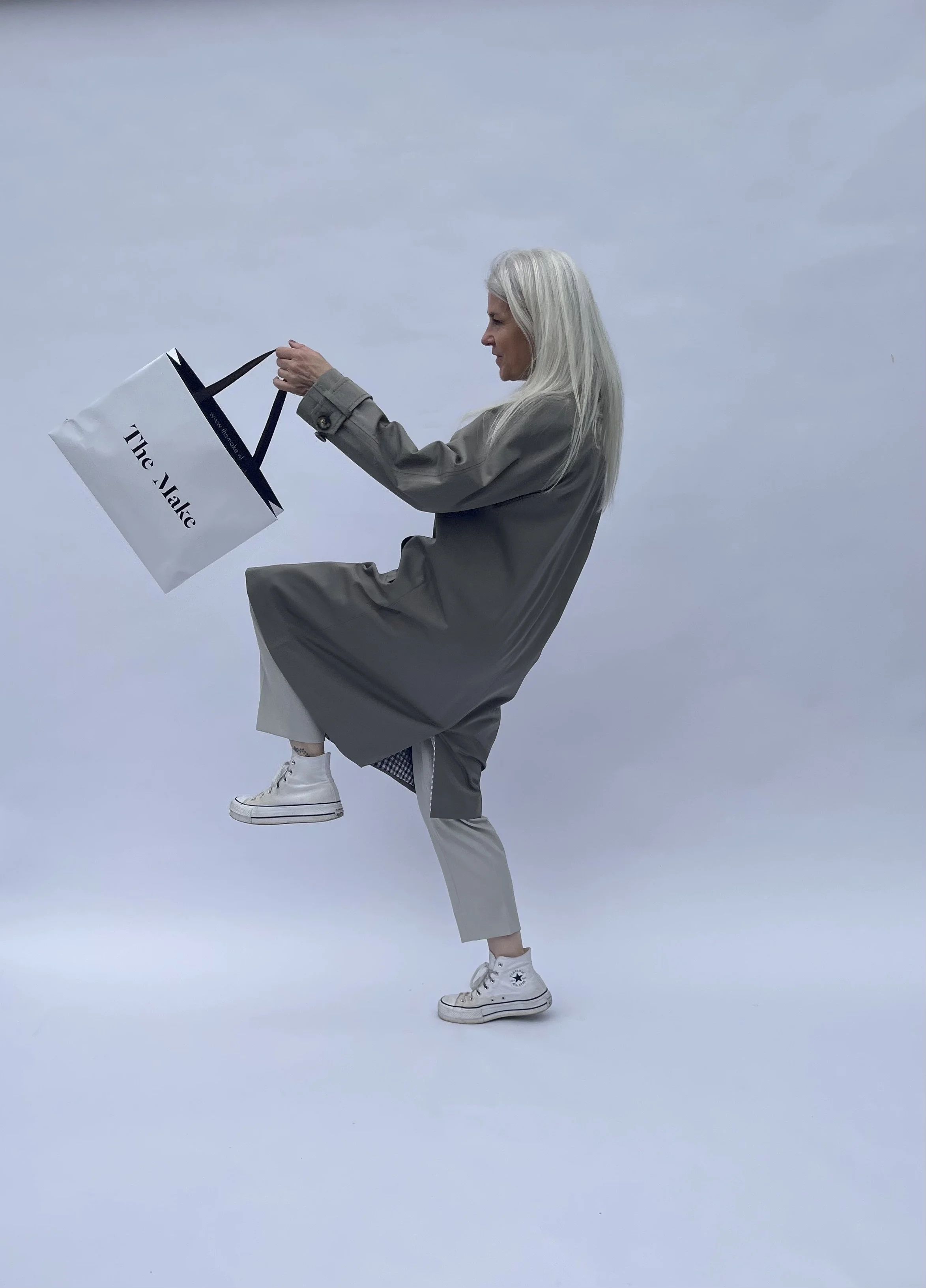 Woman with long gray hair balance on one leg while holding a shopping bag labeled 'The Alley', wearing sneakers and casual clothing, against a plain background.
