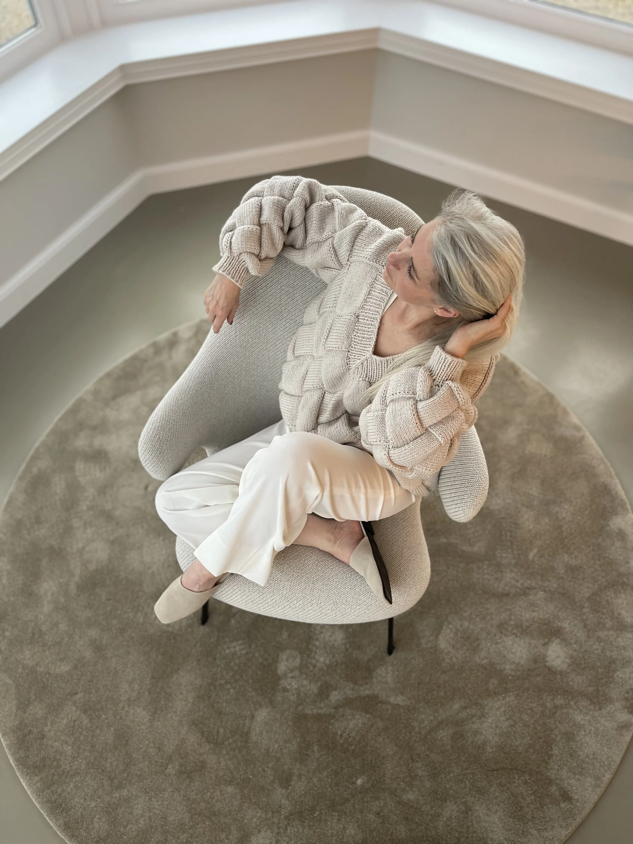 An older woman with gray hair sitting on a light-colored, modern chair on a round beige rug in a room with white walls and large windows. She is wearing a cream-colored chunky knit sweater and white pants, with her legs crossed and her head resting on her hand, looking to the side.