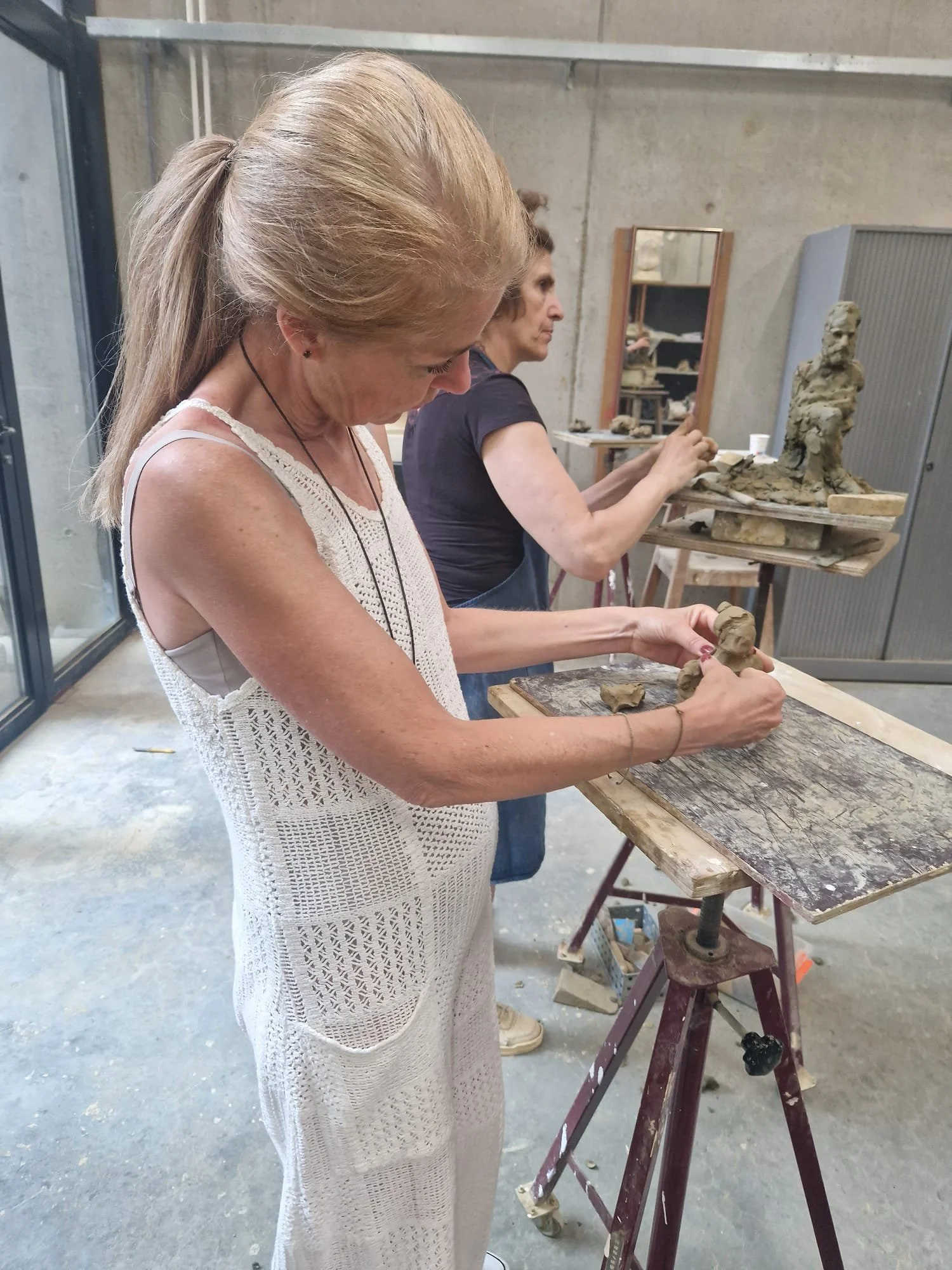 Two women are working on clay sculptures in an art studio. The woman in the foreground has blonde hair tied back and is wearing a white crochet dress, shaping clay with her hands. The woman in the background has dark hair, tied back, and is also working on a sculpture. Art supplies and sculptures are visible around them.