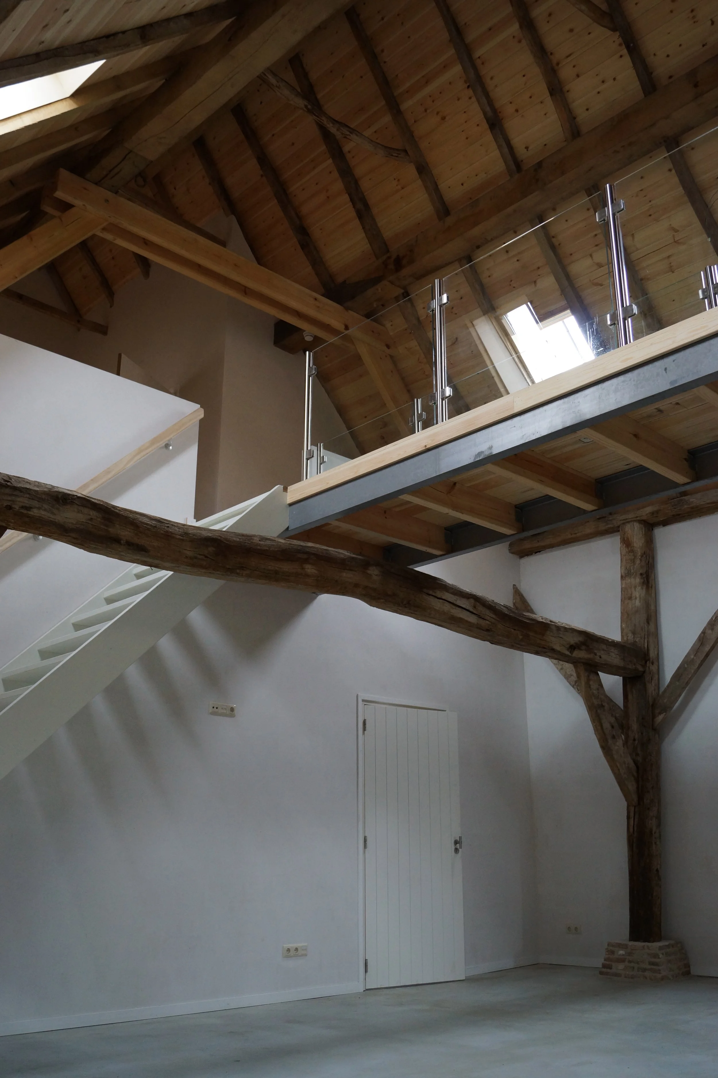 Interior view of a room featuring a wooden ceiling, a staircase, a large rustic wooden beam, and a glass railing on the upper level.
