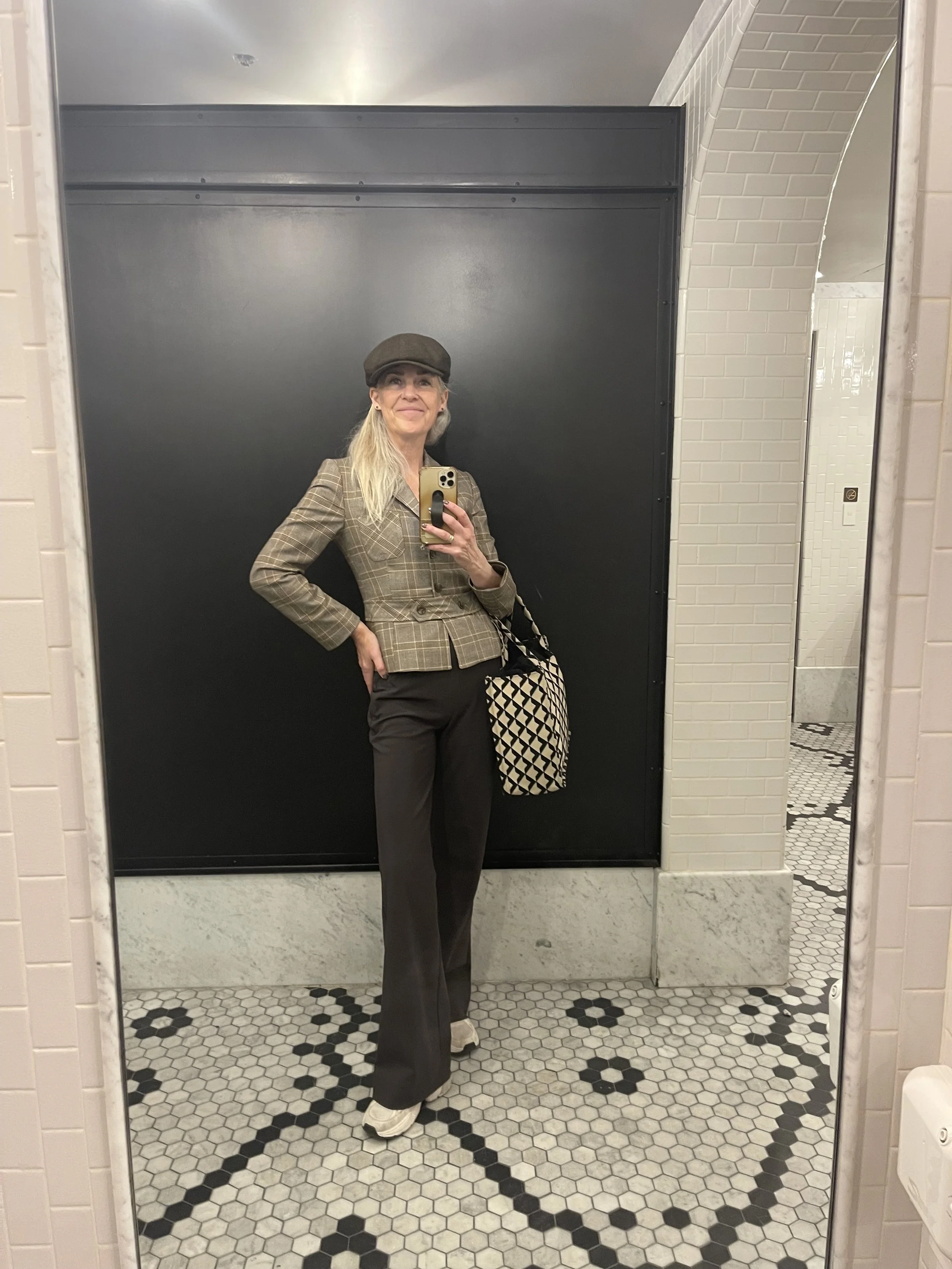 A woman taking a mirror selfie in a restroom, wearing a plaid blazer, black pants, a brown hat, and white sneakers, holding a patterned bag.
