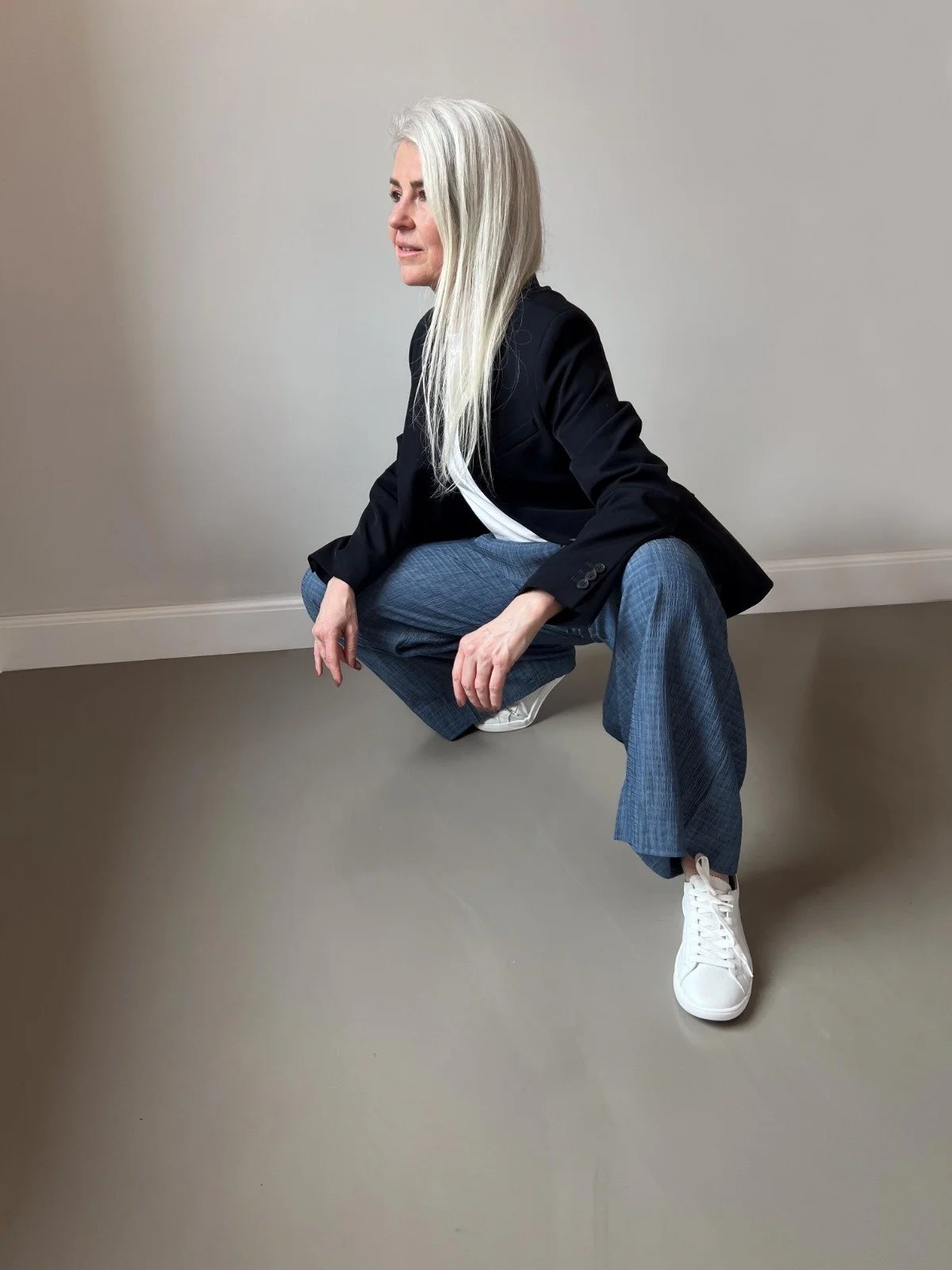 Woman with long gray hair crouching in a squatting position indoors, wearing a black blazer, gray plaid pants, and white sneakers.