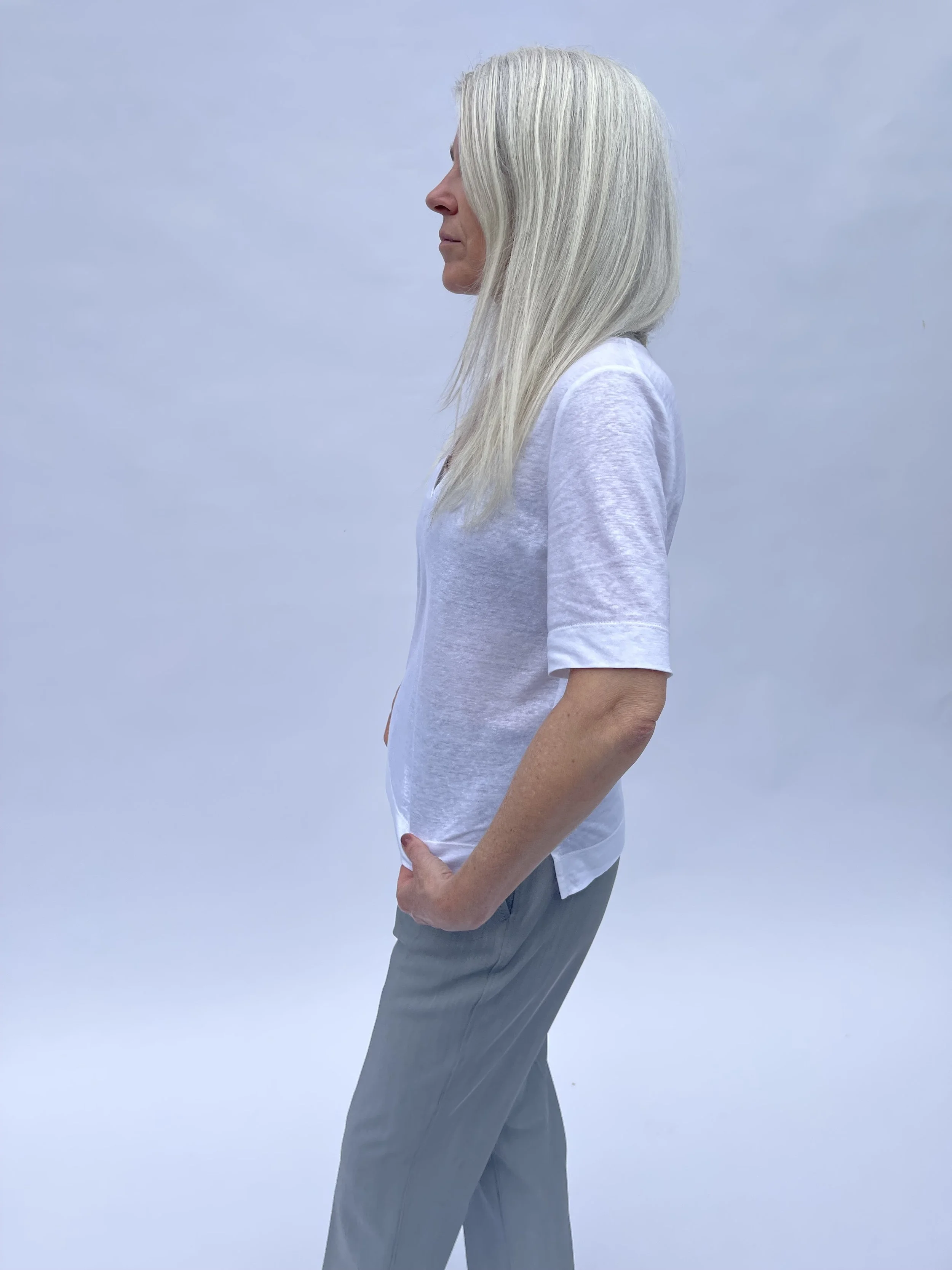 A woman with long, straight, grayish-white hair wearing a white t-shirt and gray pants standing in profile against a plain, light-colored background.