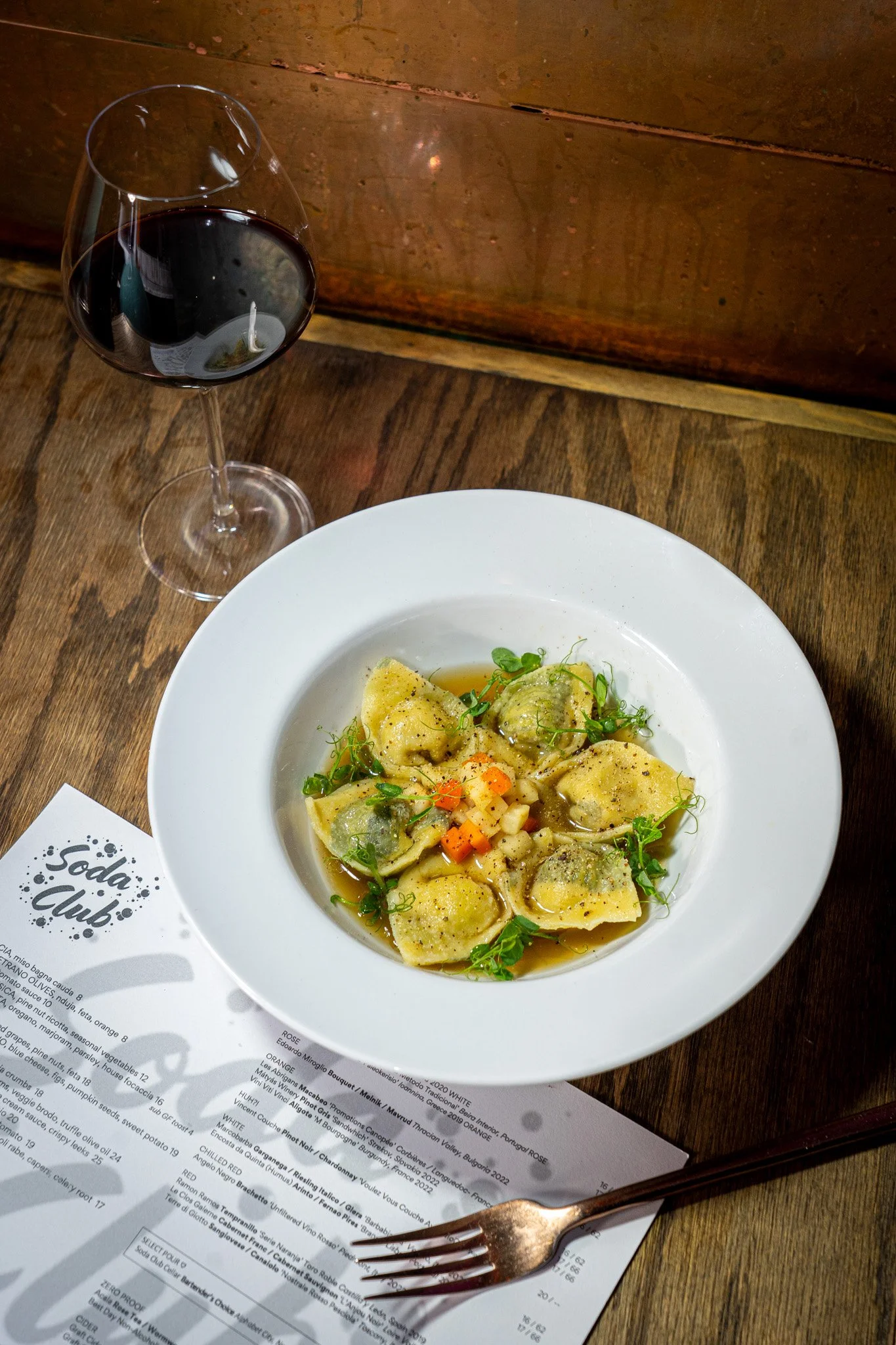 A white plate of ravioli pasta garnished with microgreens and diced vegetables, on a wooden table, with a fork, a glass of red wine, and a printed menu or wine list nearby.