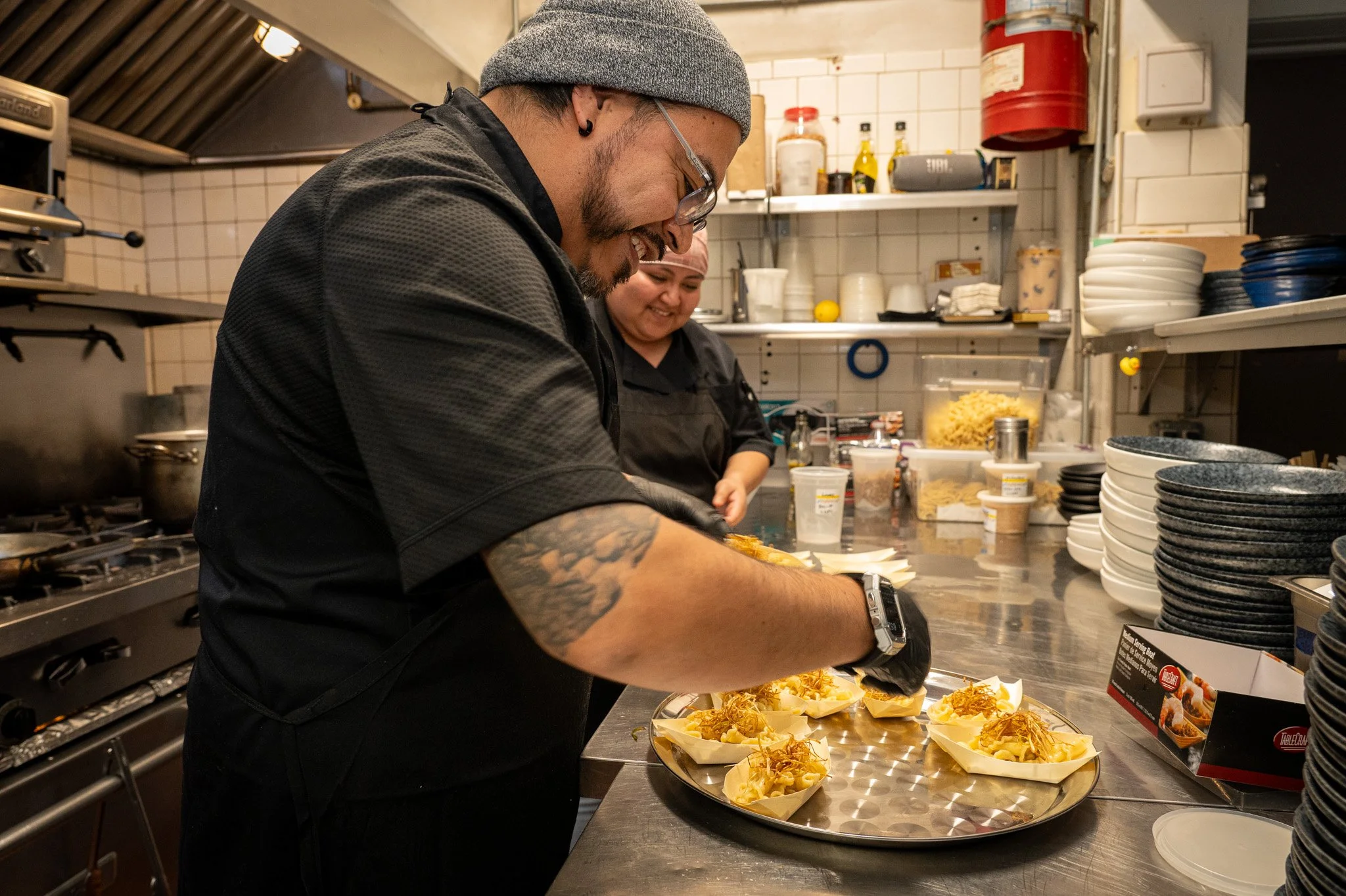 A man with tattoos on his arms and wearing a gray beanie and glasses, smiling while preparing food in a commercial kitchen. The kitchen has shelves with dishes, bottles, and containers, and a woman in the background wearing a pink head covering. The man is assembling plates with food garnished with shredded cheese and herbs.