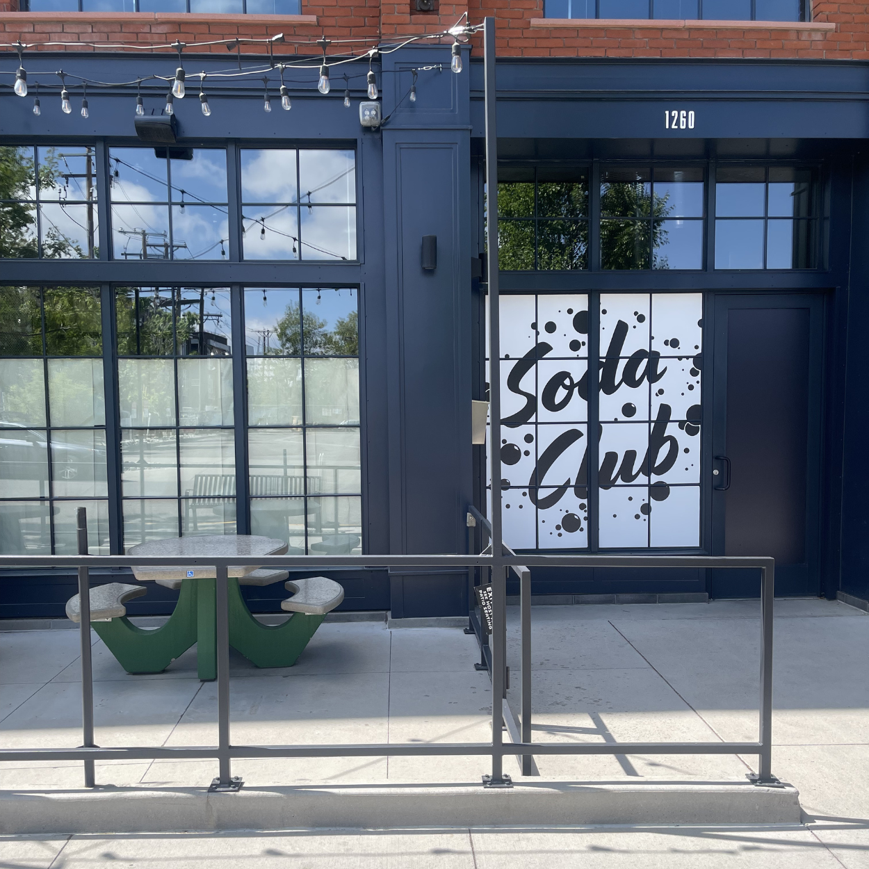 Exterior of a building with large glass windows and a door featuring the words "Sola Club" with black bubbles on a white background. A small outdoor seating area with a city sidewalk, benches, and string lights hanging above.