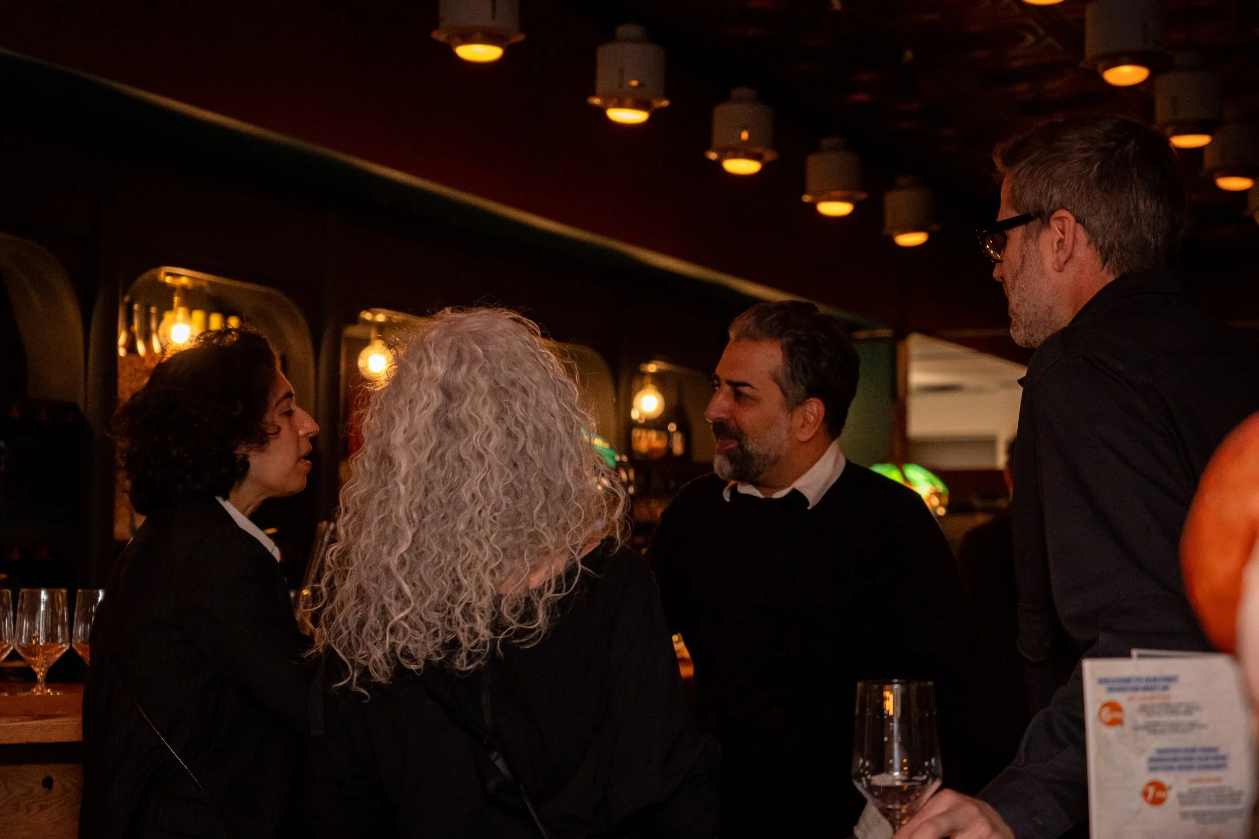 Group of four people having a conversation in a dimly lit bar, with illuminated hanging lights above and glasses of wine on the bar counter.