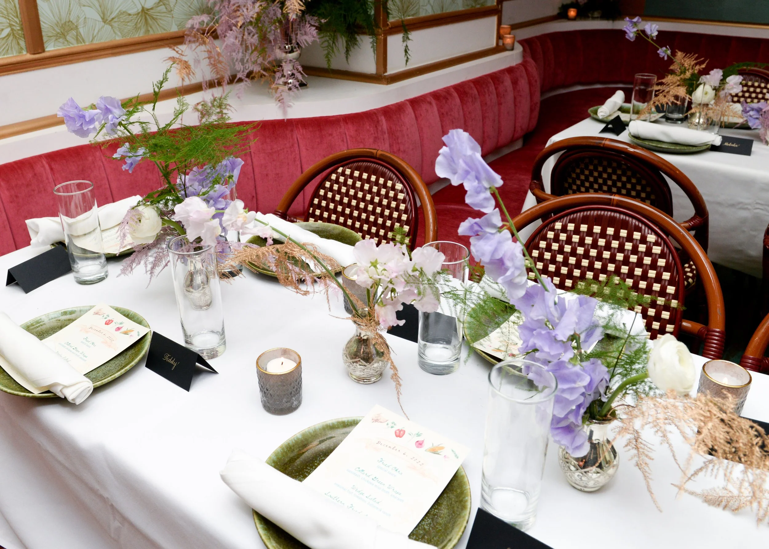 A dining table set for a meal with floral centerpieces, candles, glasses, and place settings, with a curved red upholstered banquette in the background.