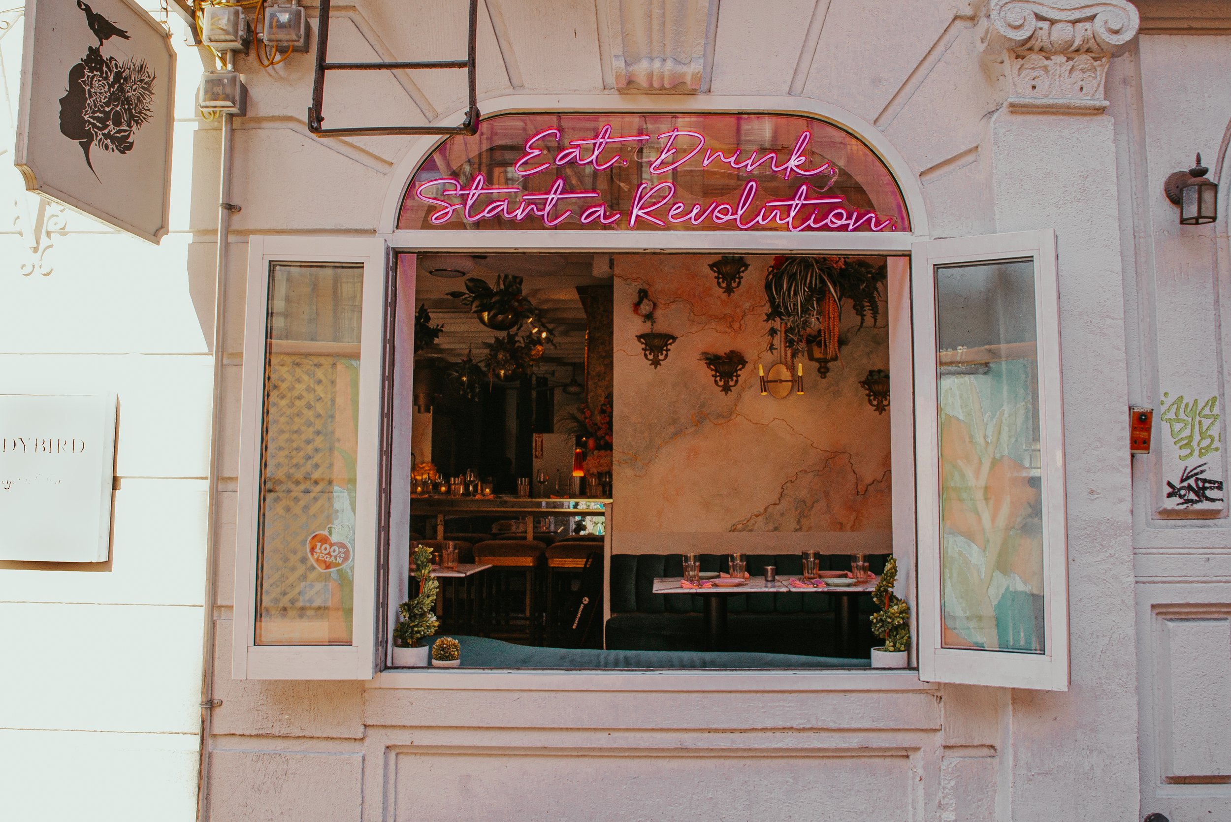 Front view of a cafe or restaurant with a pink neon sign that says 'Eat. Drink. Start a Revolution.' The establishment has open double doors, with interior decor including plants, hanging lights, and framed art on the walls.