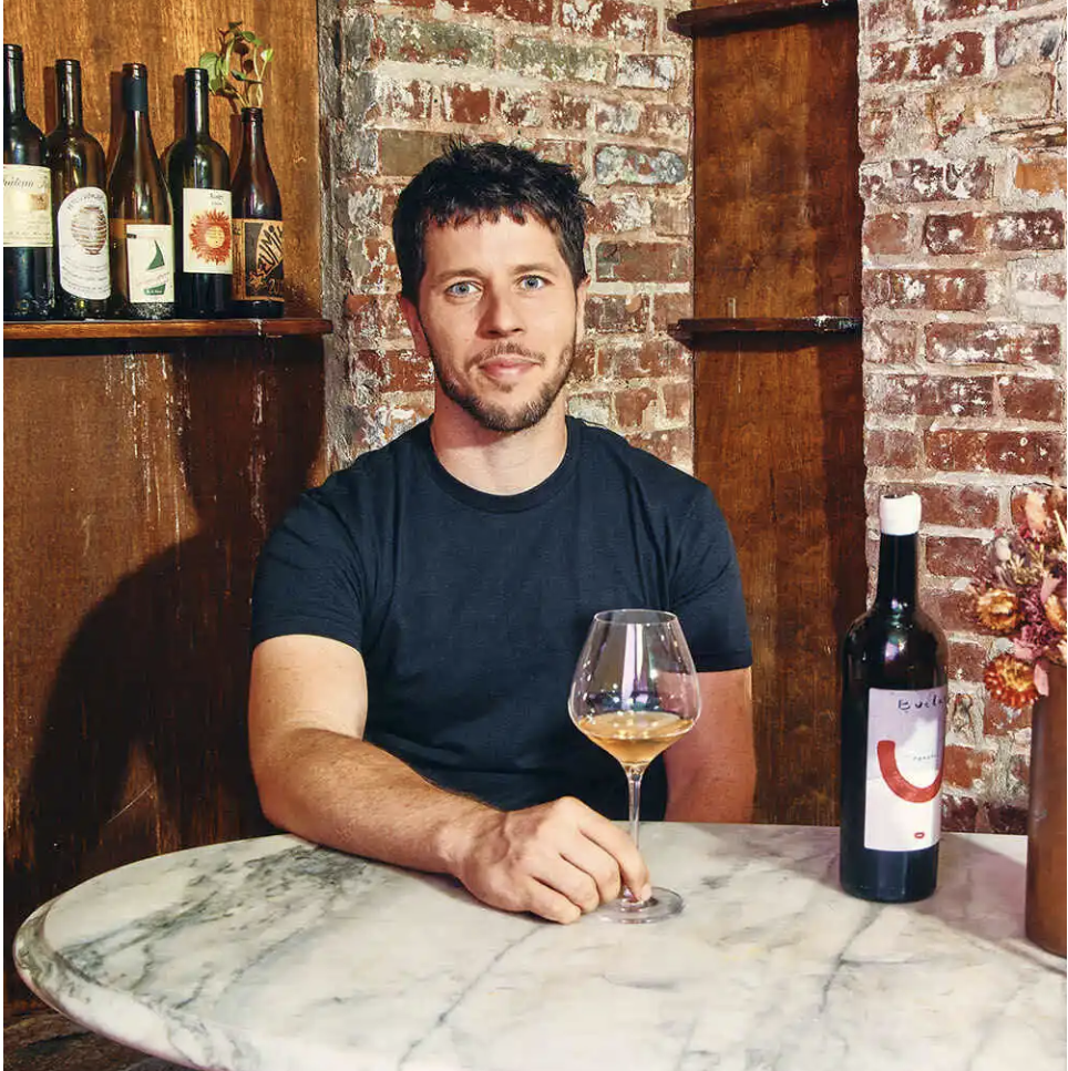 A man with dark hair and a beard, wearing a black t-shirt, sitting at a marble table in a rustic setting, holding a glass of white wine. Behind him are brick walls, wooden shelves with wine bottles, and a flower arrangement in a vase.