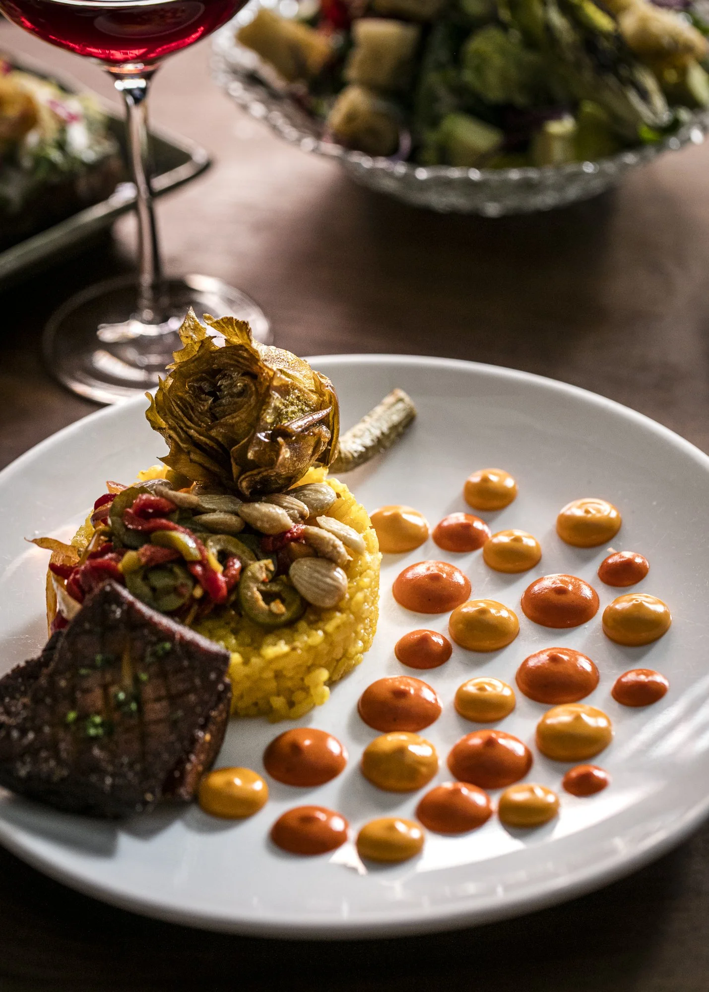 Close-up of a gourmet plated dish with a small roasted artichoke on top, accompanied by dots of orange sauce on a white plate, with a glass of red wine and a bowl of salad in the blurred background.