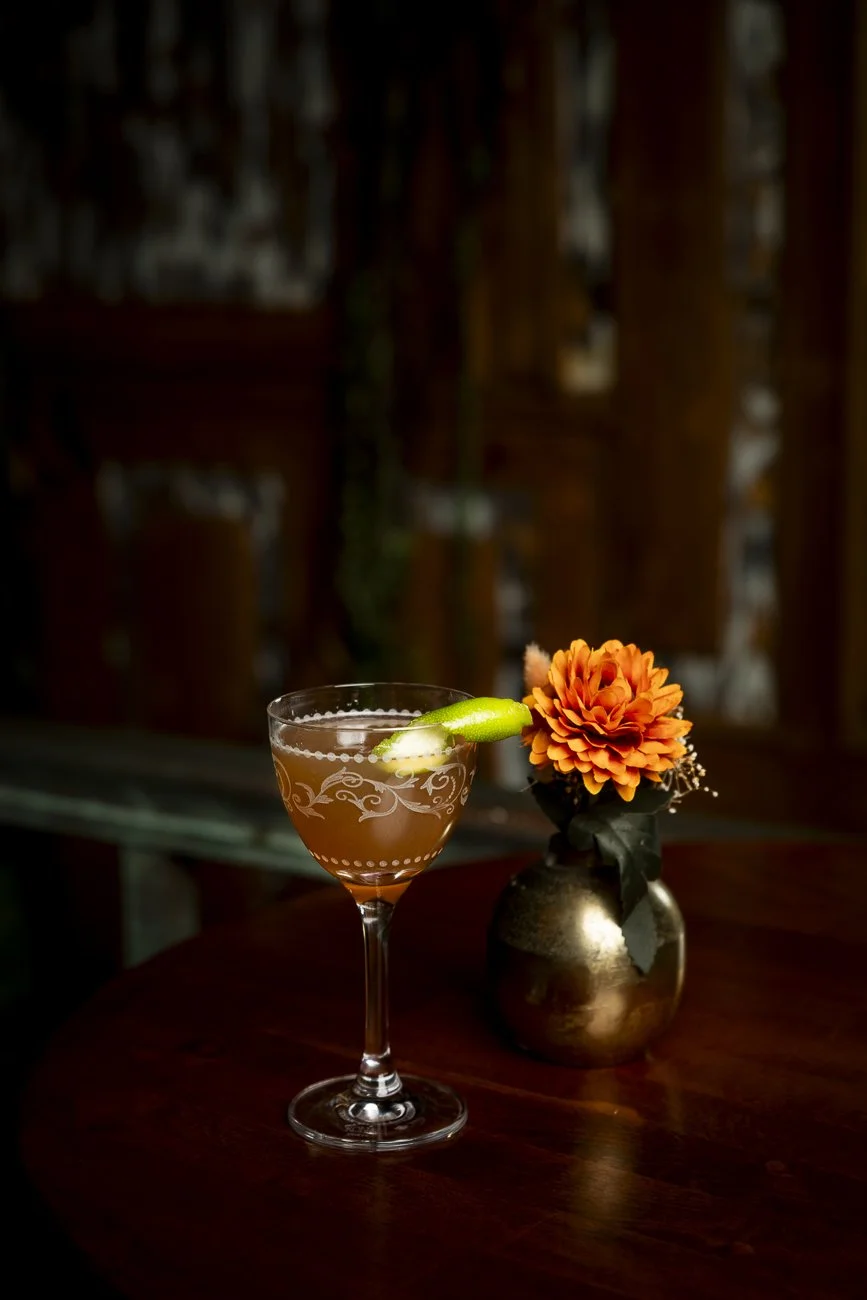 Cocktail on a wooden table with an orange flower in a brass vase, dark background.