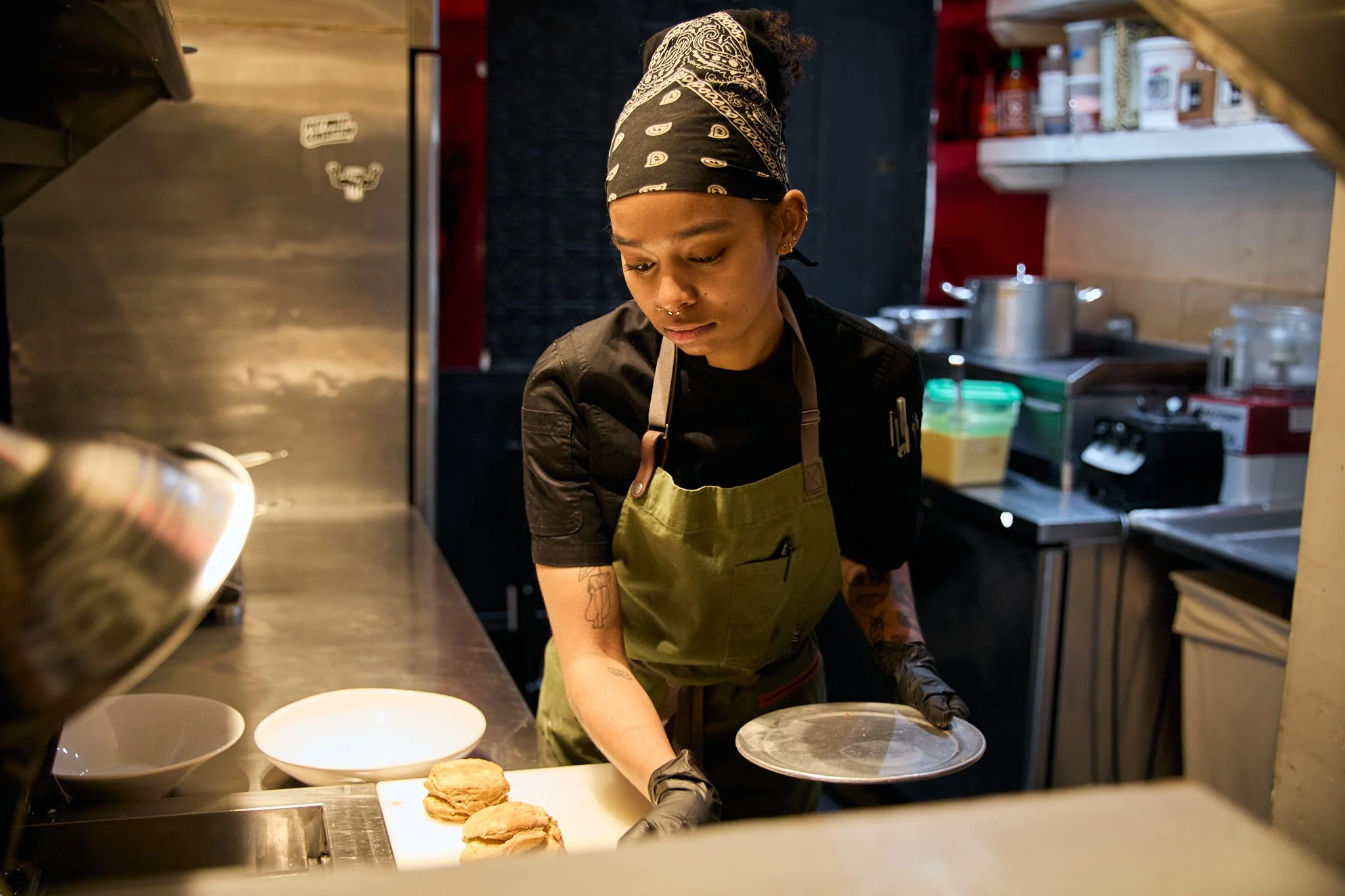 A young woman with tattoos and dark hair tied back, wearing a bandana and black shirt, is working in a kitchen. She is placing what appears to be flatbread or similar food onto a surface, with bowls and kitchen equipment visible around her.