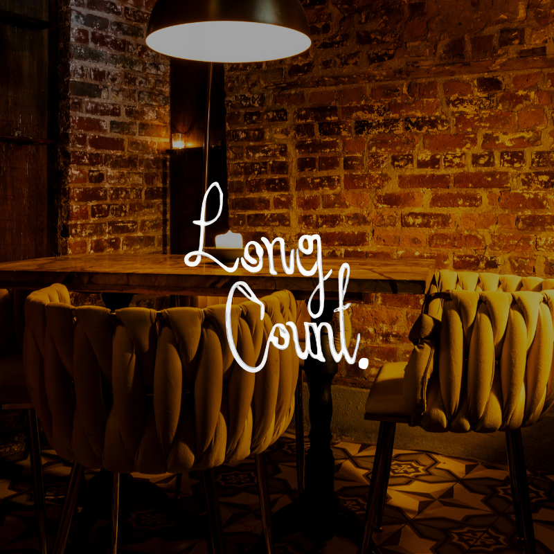 Cozy dining space with exposed brick wall, a hanging lamp, a wooden table, and plush beige chairs. "Loving Count" sign in white script on the wall.