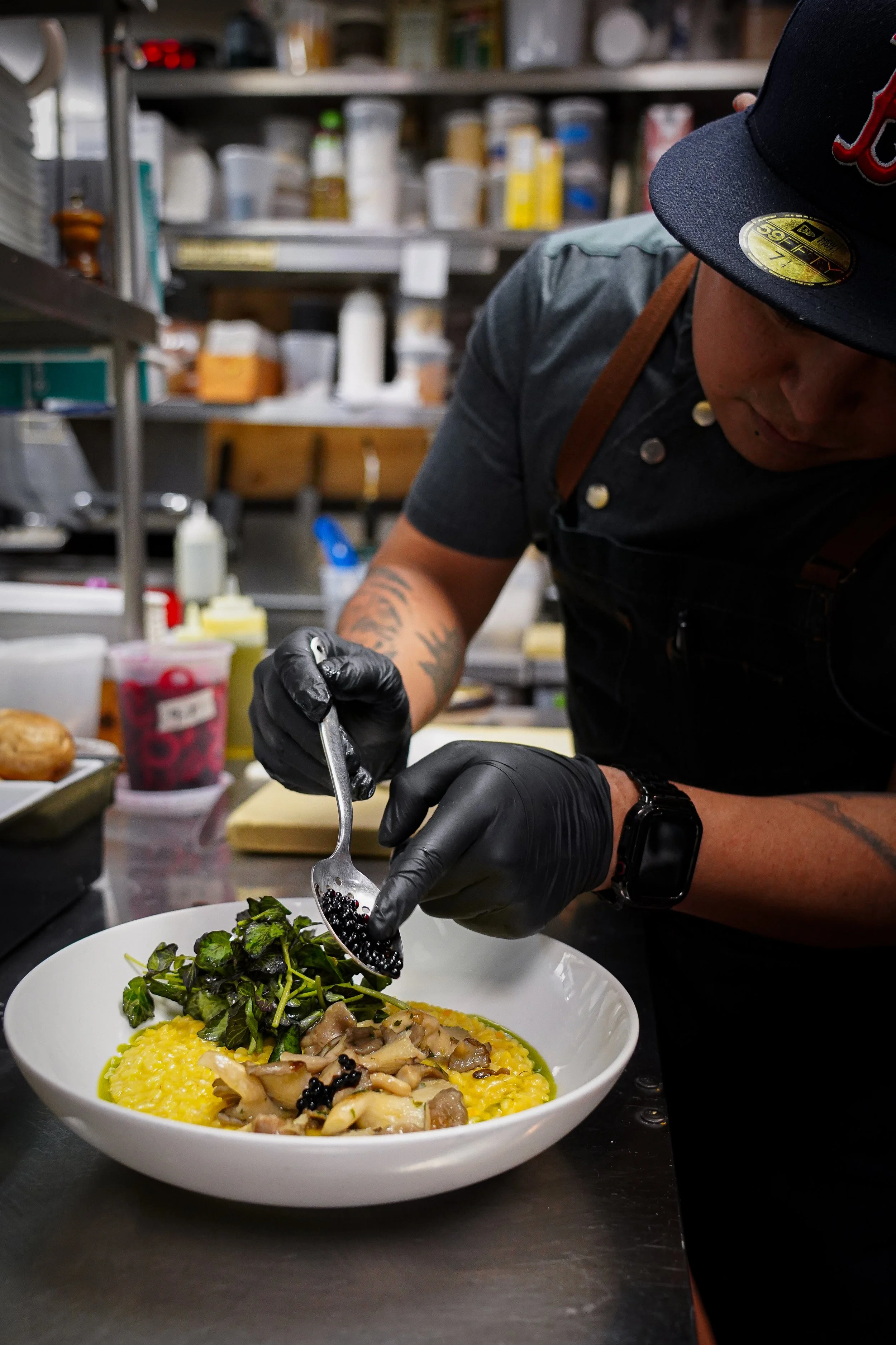 A person wearing black gloves and a wristwatch is garnishing a bowl of yellow risotto with mushrooms, greens, and blackberries in a professional kitchen with shelves of spices and ingredients in the background.