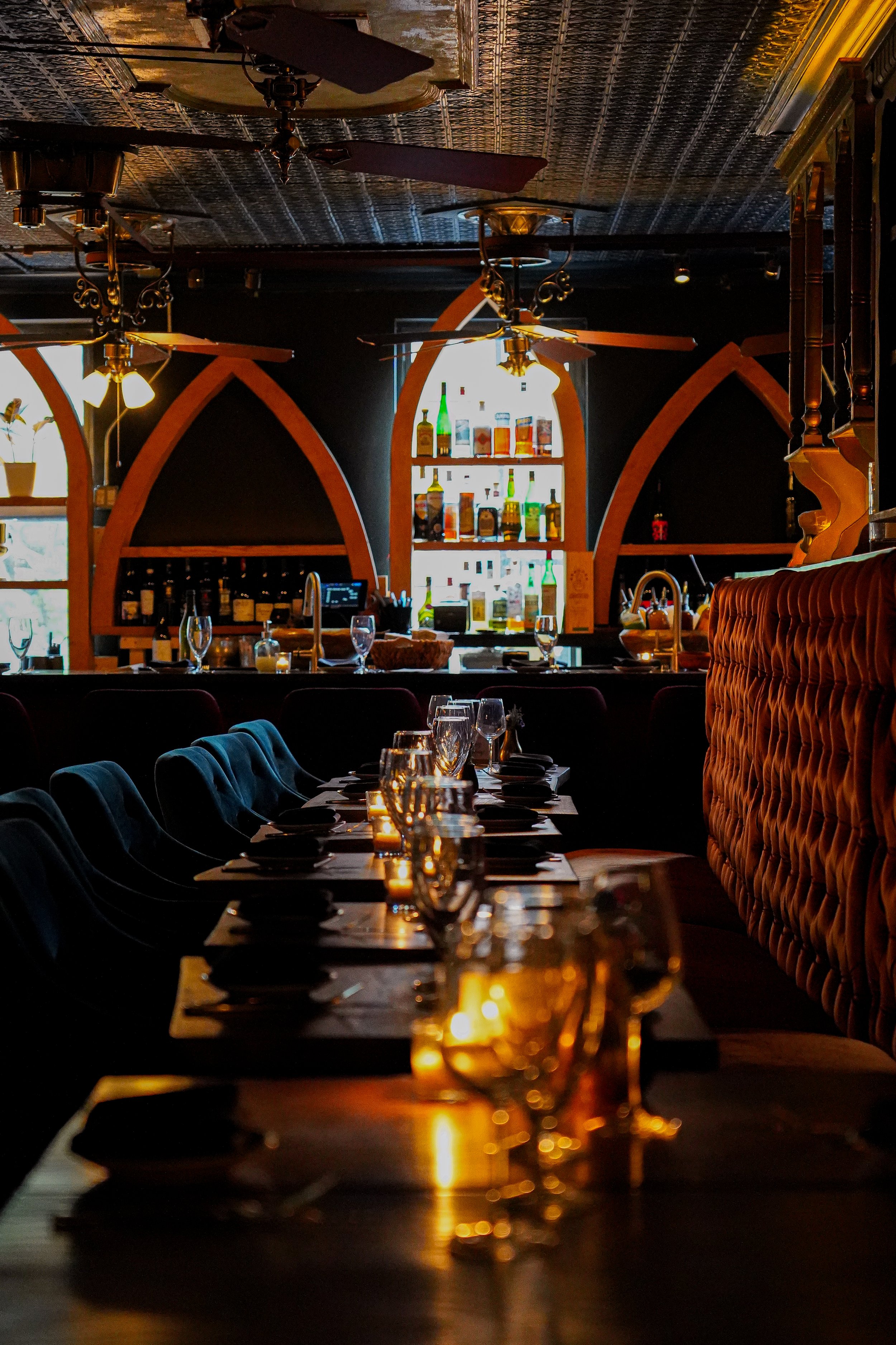 Elegant restaurant interior with a long dining table set with black napkins, wine glasses, and a candle in the foreground. In the background, a bar area with a lit archway displaying bottles of liquor and barware, and decorative ceiling fans overhead.