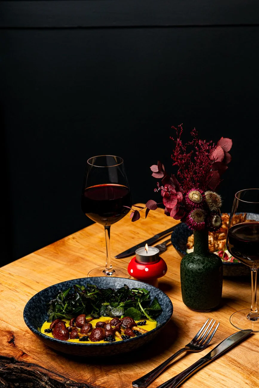 A dining table set with two glasses of red wine, a small lit candle in a red holder, a blue bowl with a salad, and a vase of dark pink flowers against a black background.