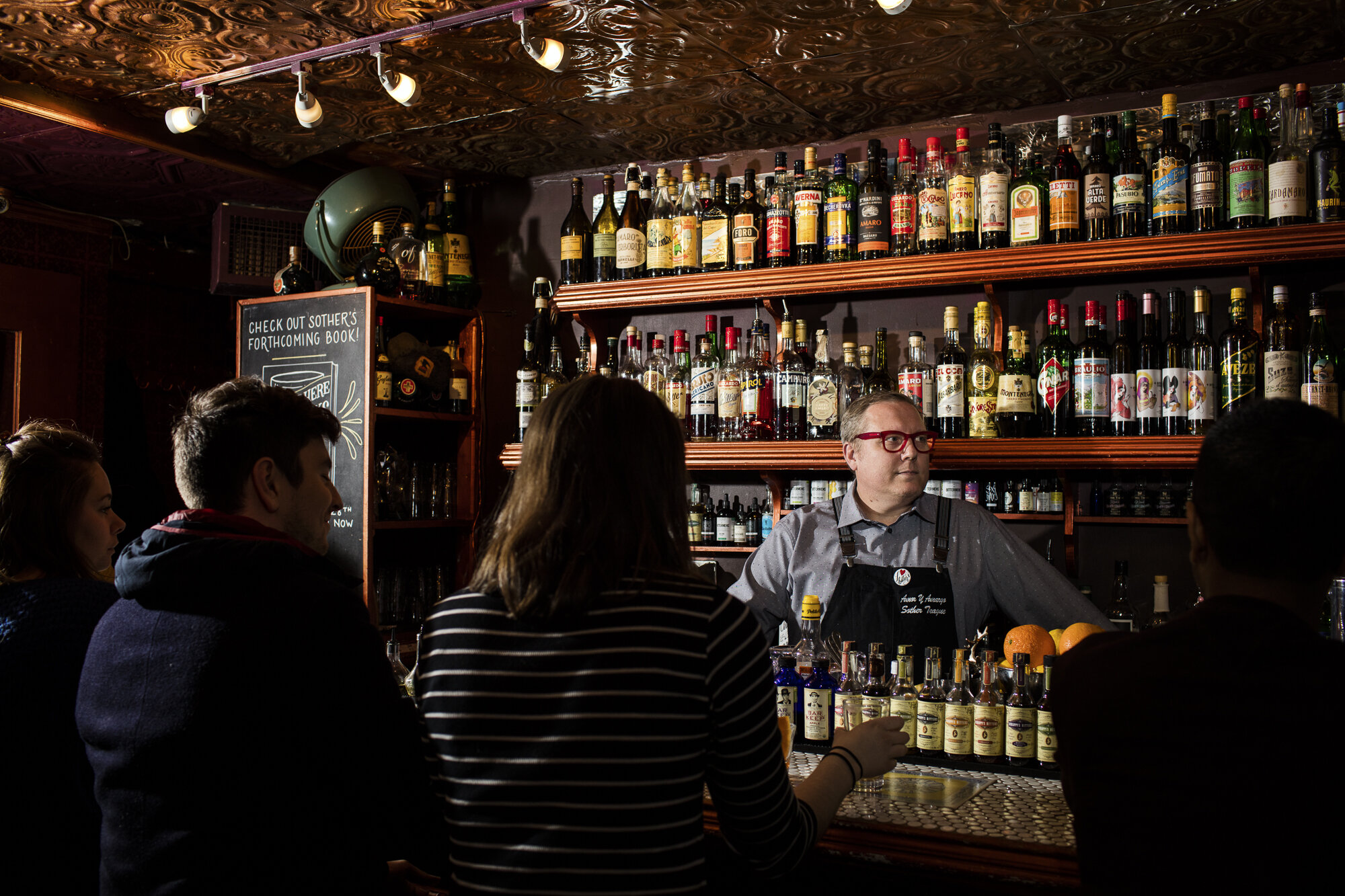 A bartender behind a bar with various liquor bottles on shelves, serving customers in a dimly lit bar or pub with a copper patterned ceiling and a chalkboard sign.