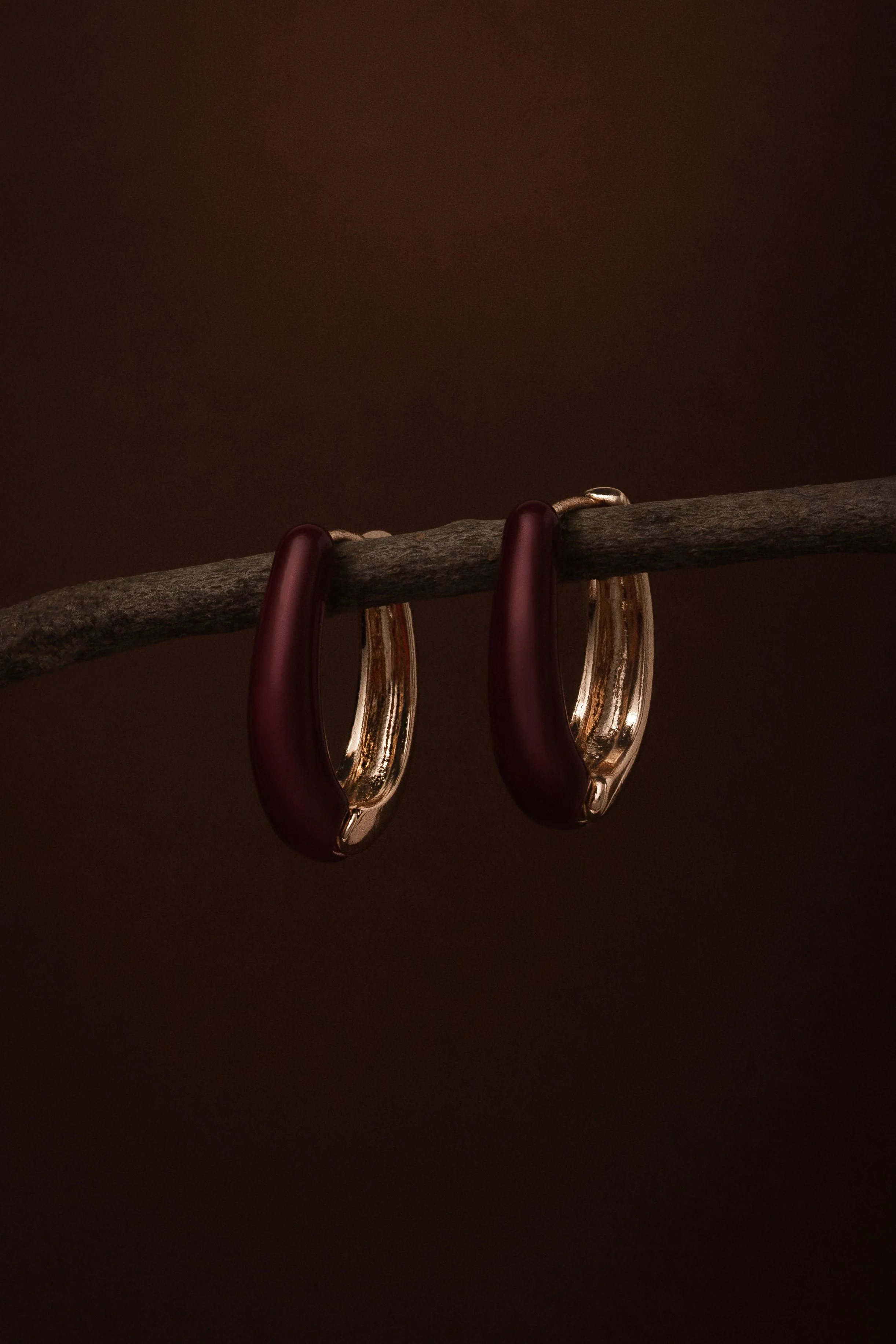 A pair of earrings hang from a textured wooden stick against a dark brown background.