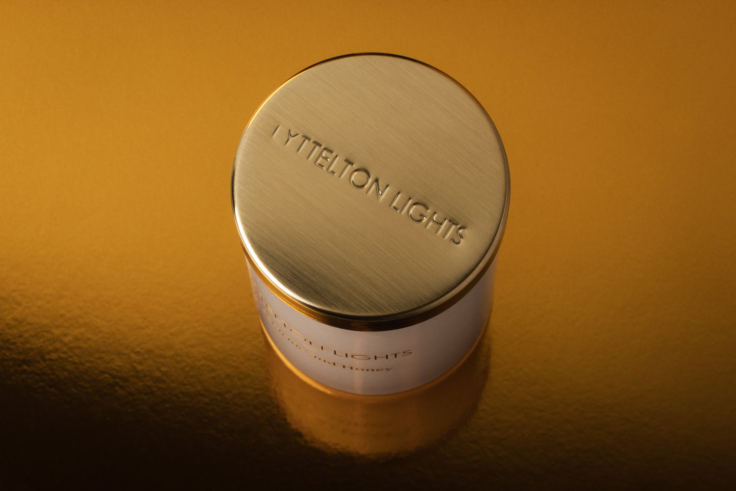 Close-up of a metallic lid with the words 'YIT ELTON LIGHTS' engraved, set on a cylindrical container, against a golden textured background.