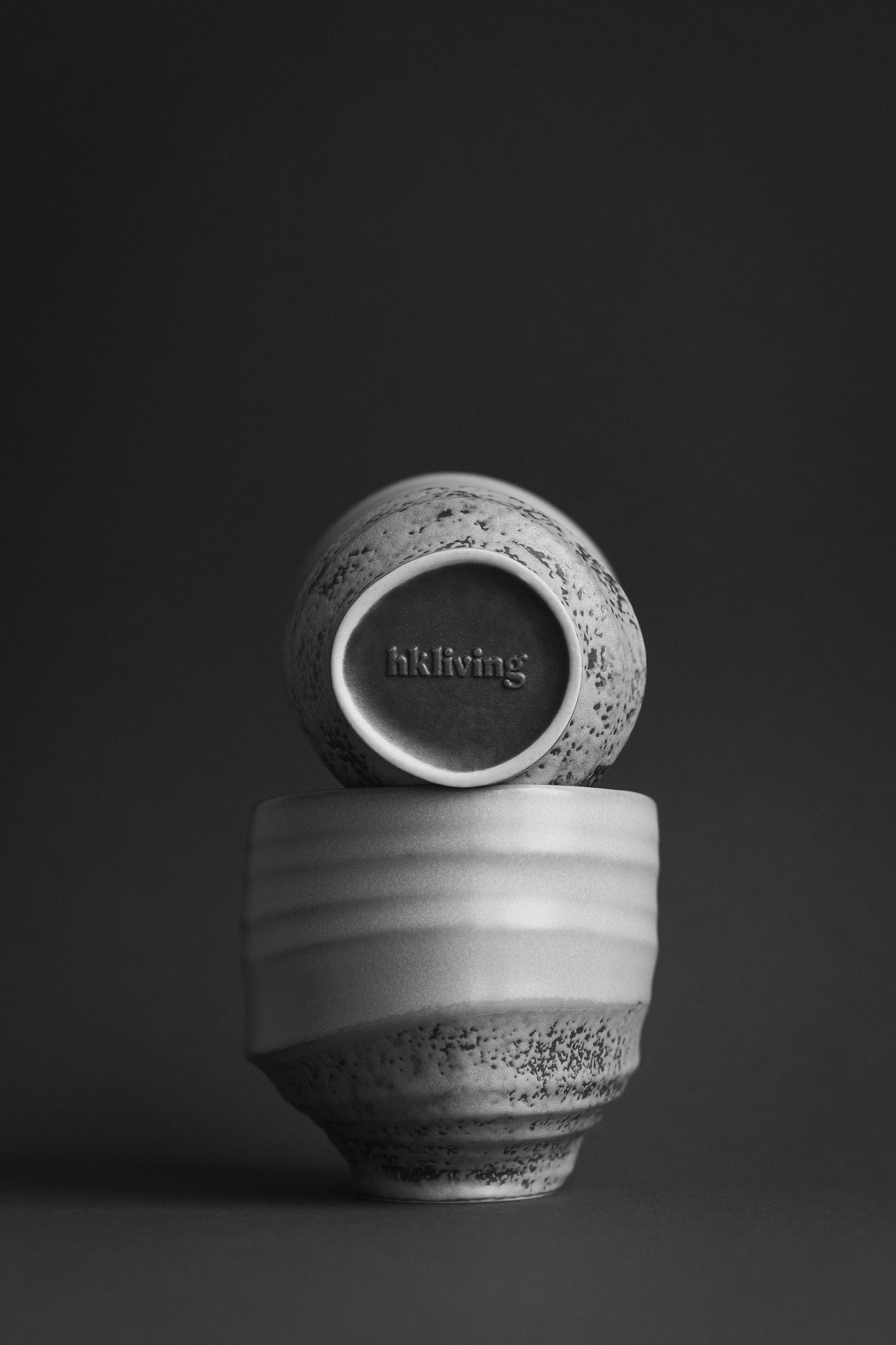 A black and white photograph of a ceramic cup with the word 'hiking' inscribed inside. The cup is placed upside down on a textured, rounded base, with a plain, dark background.