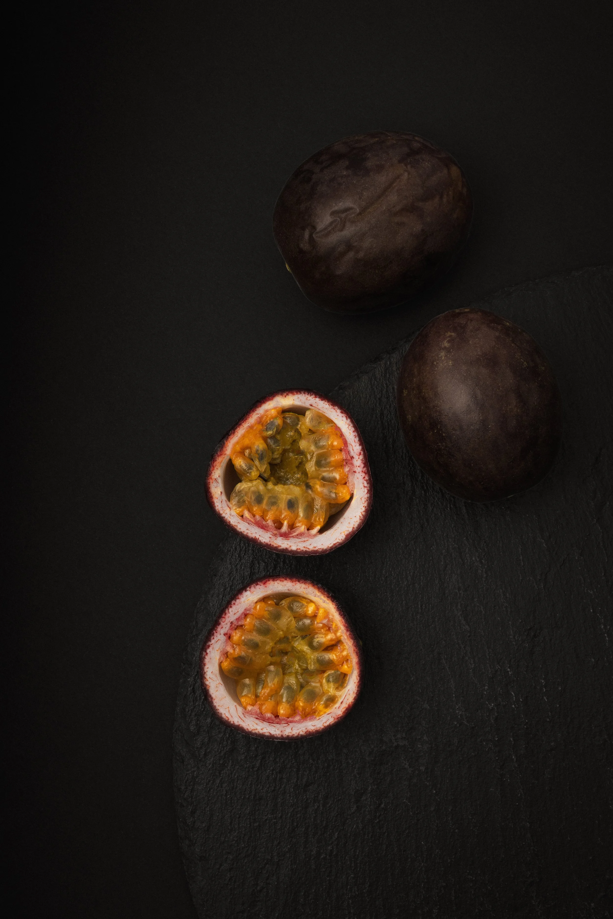 Halved passion fruits with yellow pulp and black seeds, placed on a black textured surface.