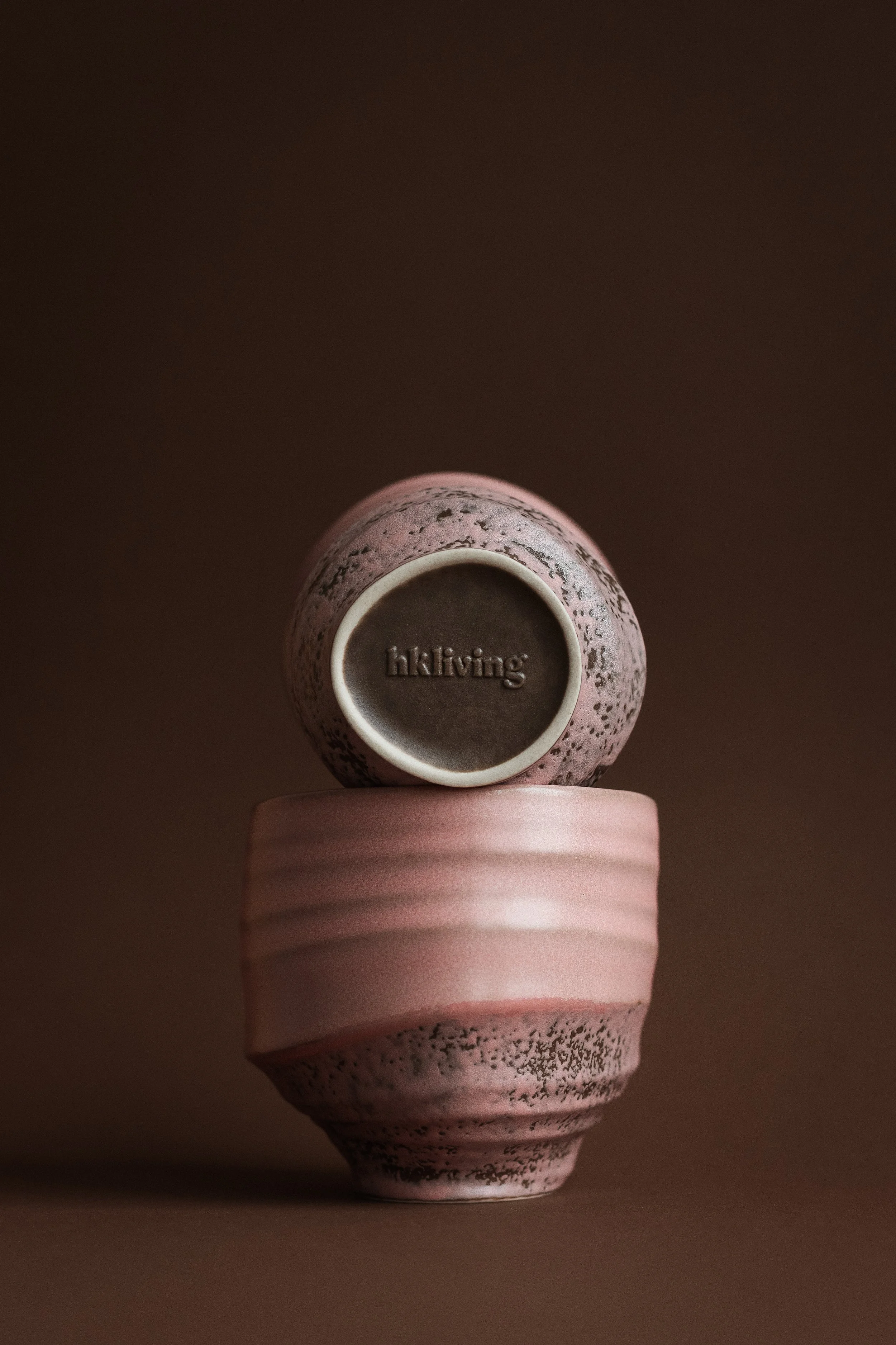 Top view of a pink ceramic cup, showing the bottom with the embossed brand name 'hkliving', resting on a pink saucer with a textured striped pattern. The background is plain and dark.