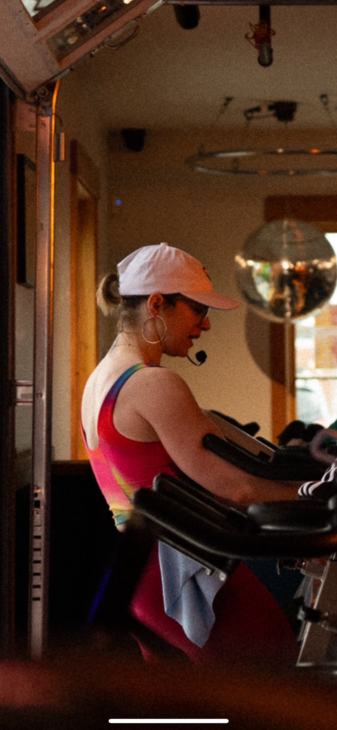 A woman with dark hair in a high bun, wearing a Nike sports bra and patterned leggings, holding dumbbells, standing against a plain background.
