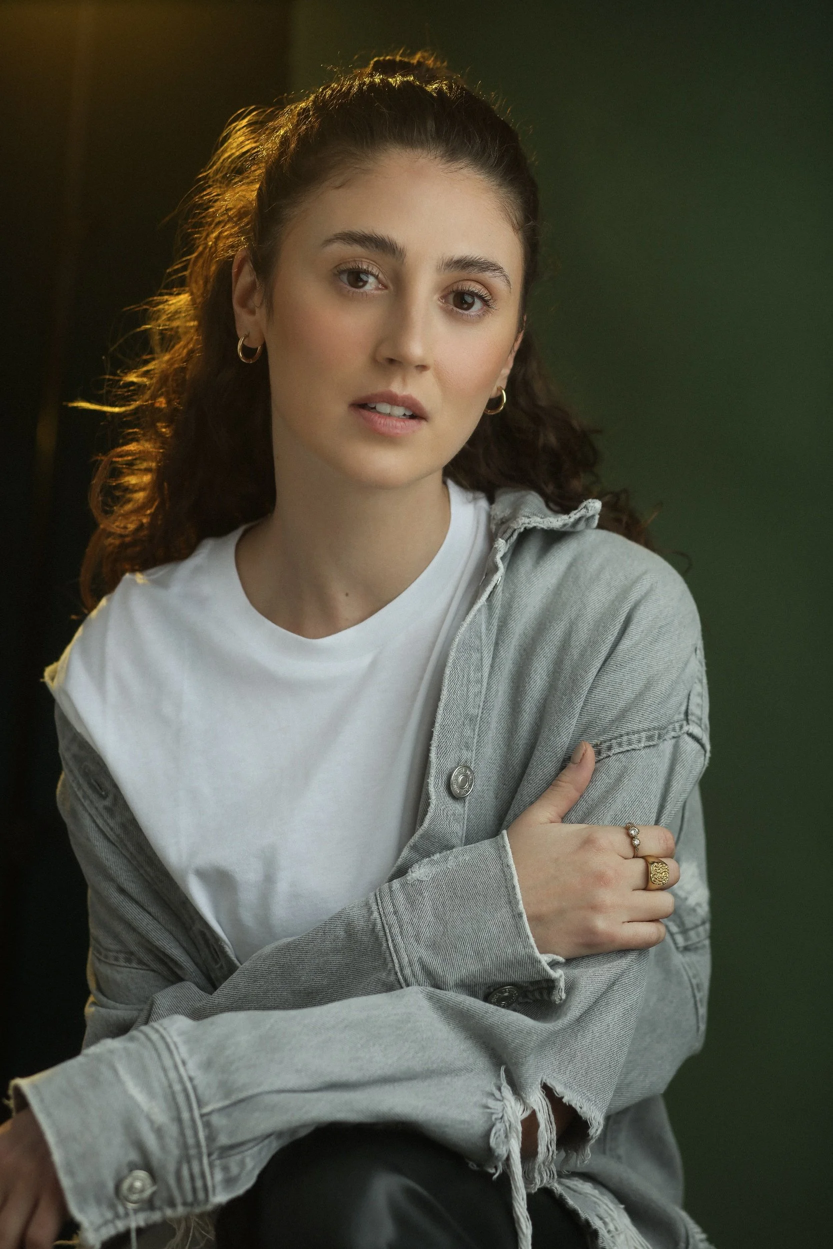 Woman with curly hair wearing a denim jacket and white t-shirt, posing against a dark green background.