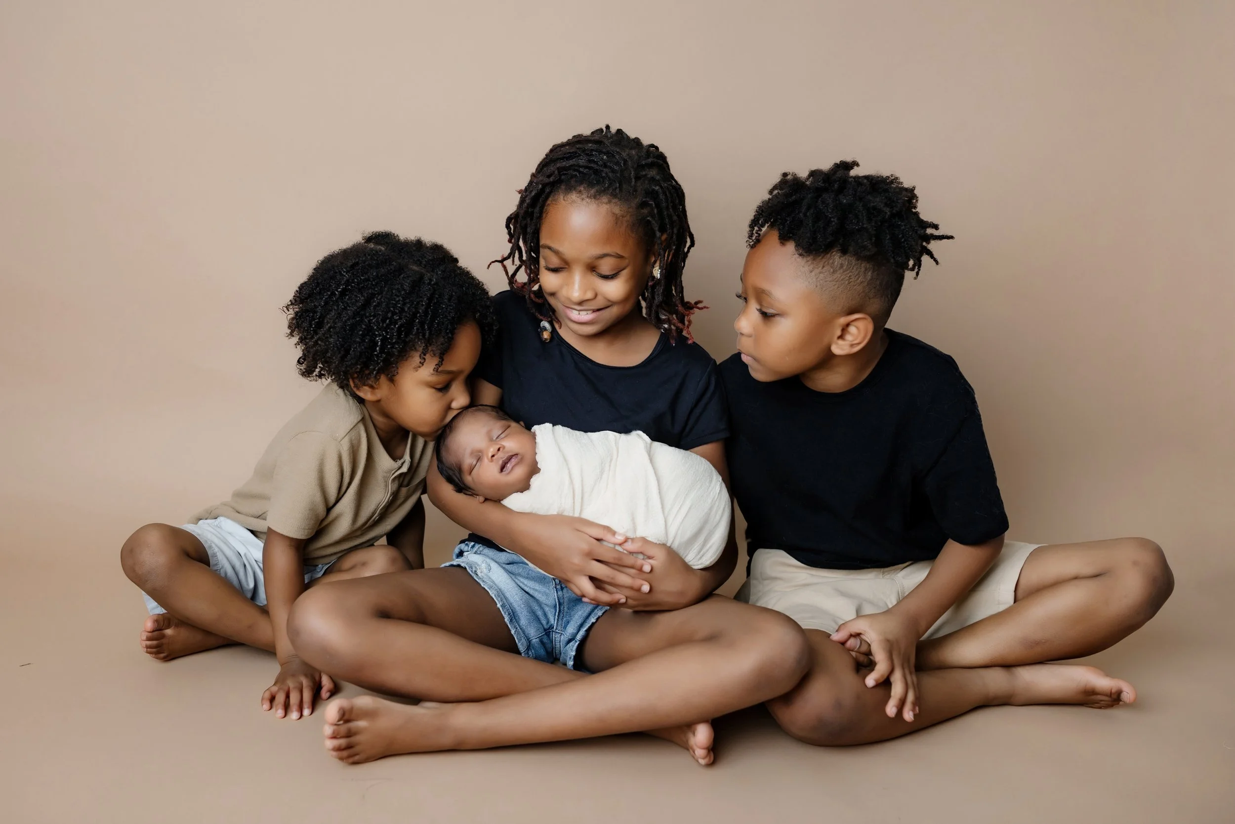 Four children, three older siblings and a newborn, sitting closely together on a plain beige background. The eldest girl, with dreadlocks, gently holds the newborn wrapped in a white blanket while the other two kids, a boy and girl, look affectionate