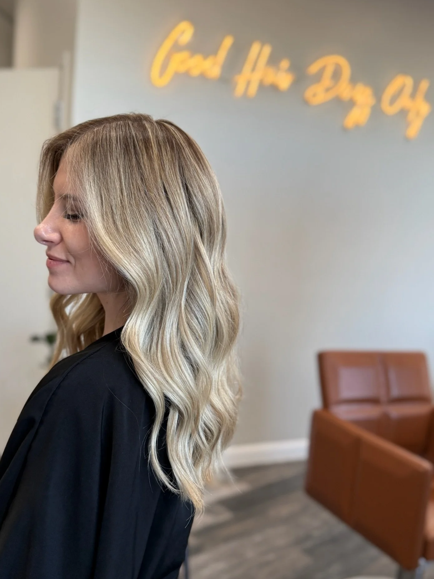 Side profile of a woman with long, wavy blonde hair, seated in a salon. In the background, a neon sign reads 'Good Hair Day QA' on the wall.