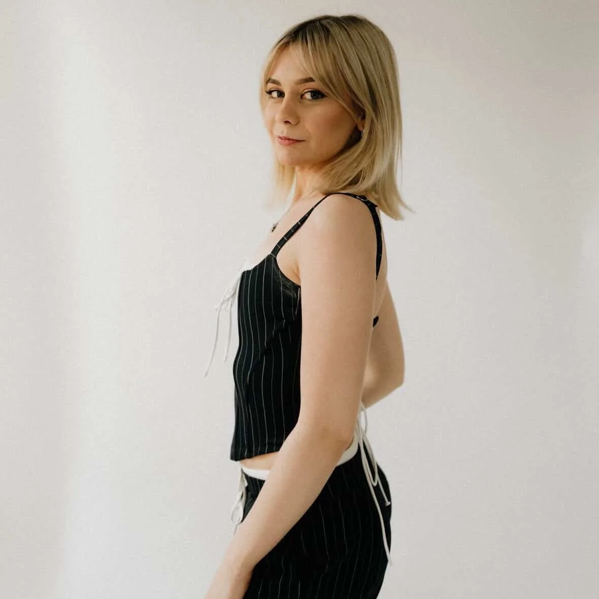 A woman with blonde hair wearing a black pinstriped tank top and matching pants, standing against a plain white wall, looking over her shoulder with a slight smile.
