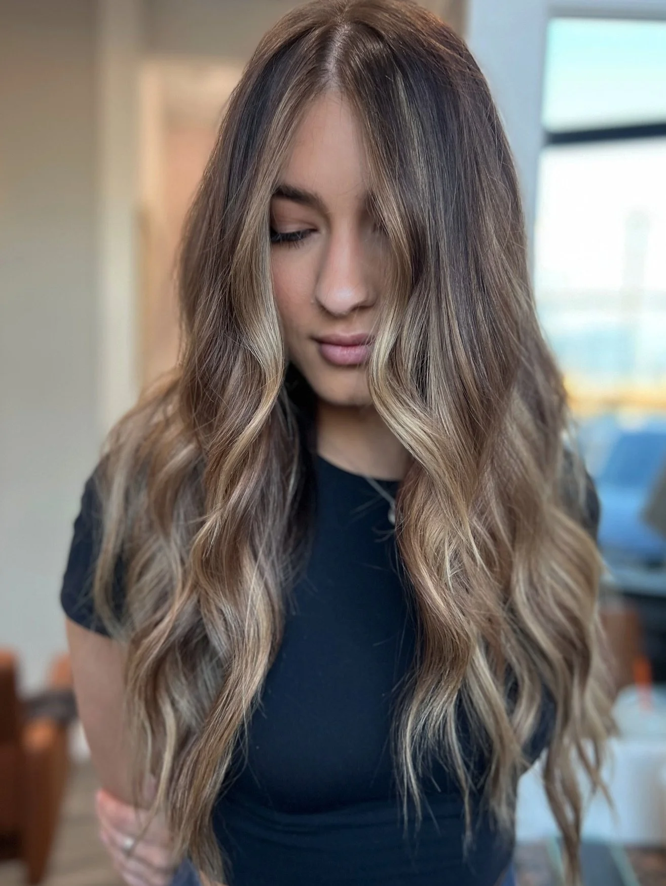 Portrait of a young woman with long wavy brown hair, wearing a black top, looking downward with her eyes closed, indoors.