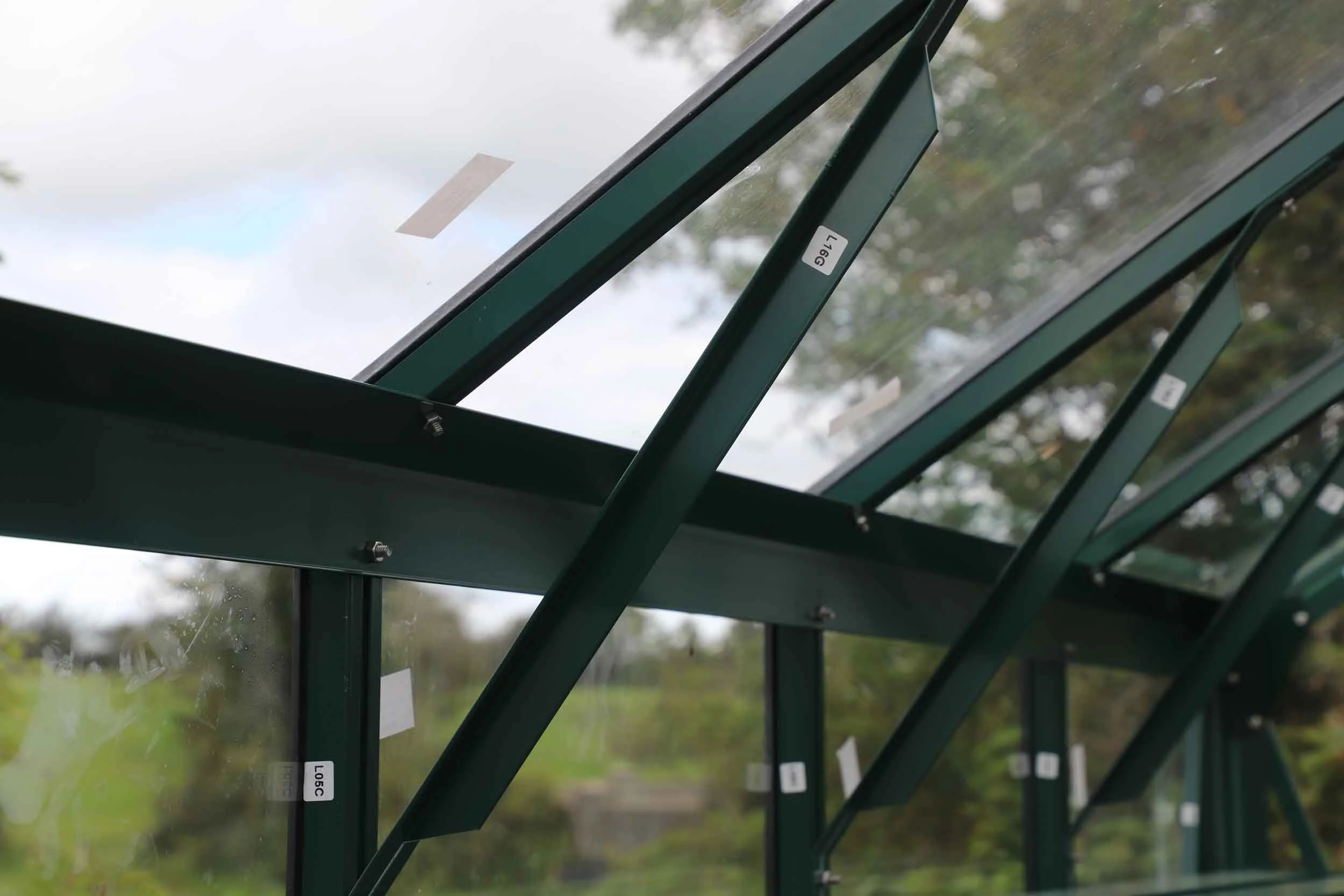 Multiple green metal and glass greenhouse panels tilted open with labels and latches visible, set against a cloudy sky and trees in the background.