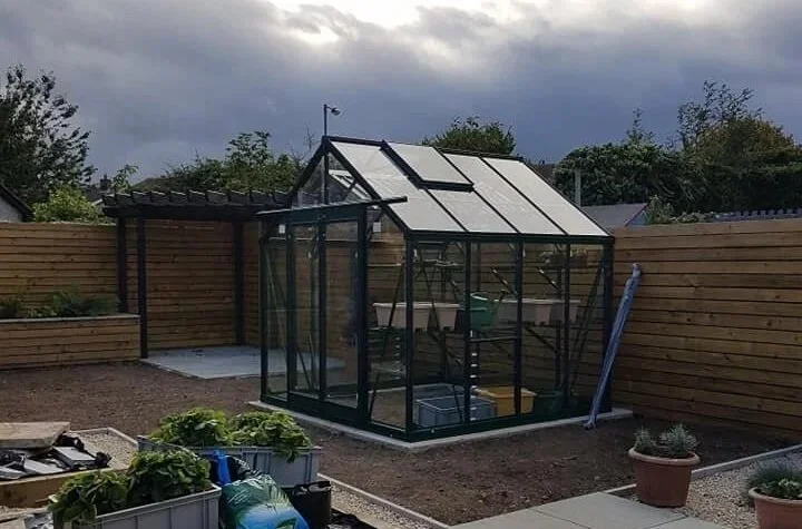 A small greenhouse in a backyard surrounded by a wooden fence. There are potted plants and garden tools nearby, with cloudy skies overhead.
