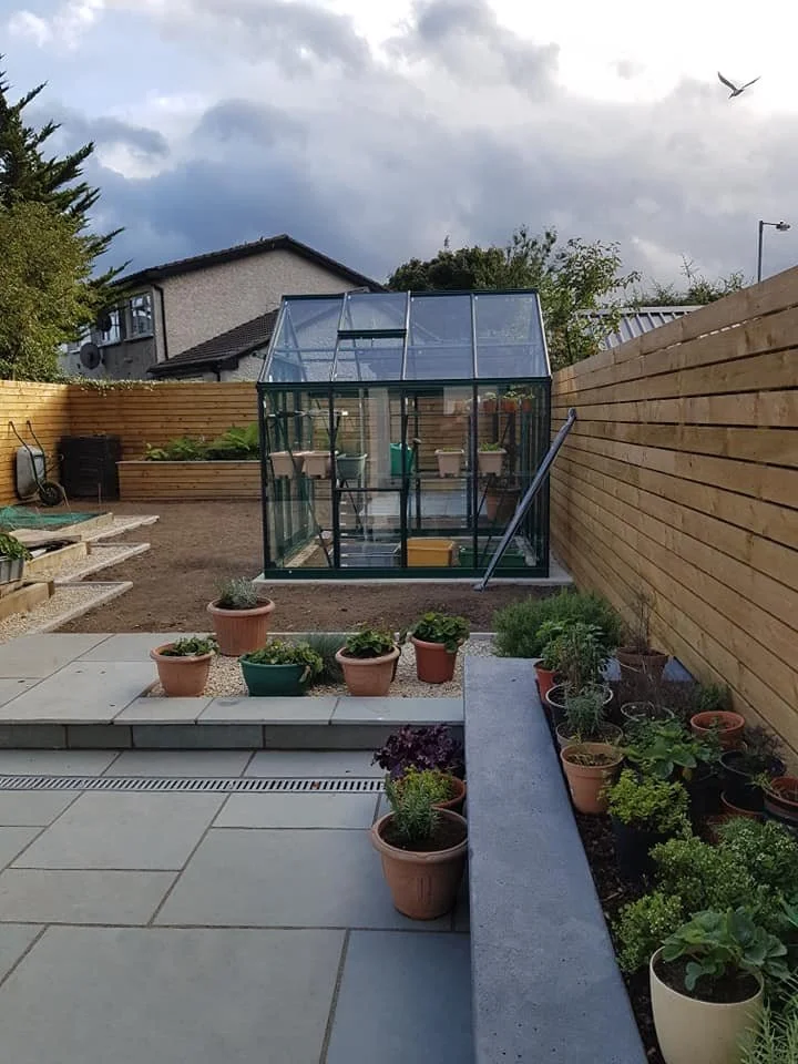 A backyard garden with a small 8x8 ft greenhouse, potted plants, and a wooden fence under a cloudy sky.