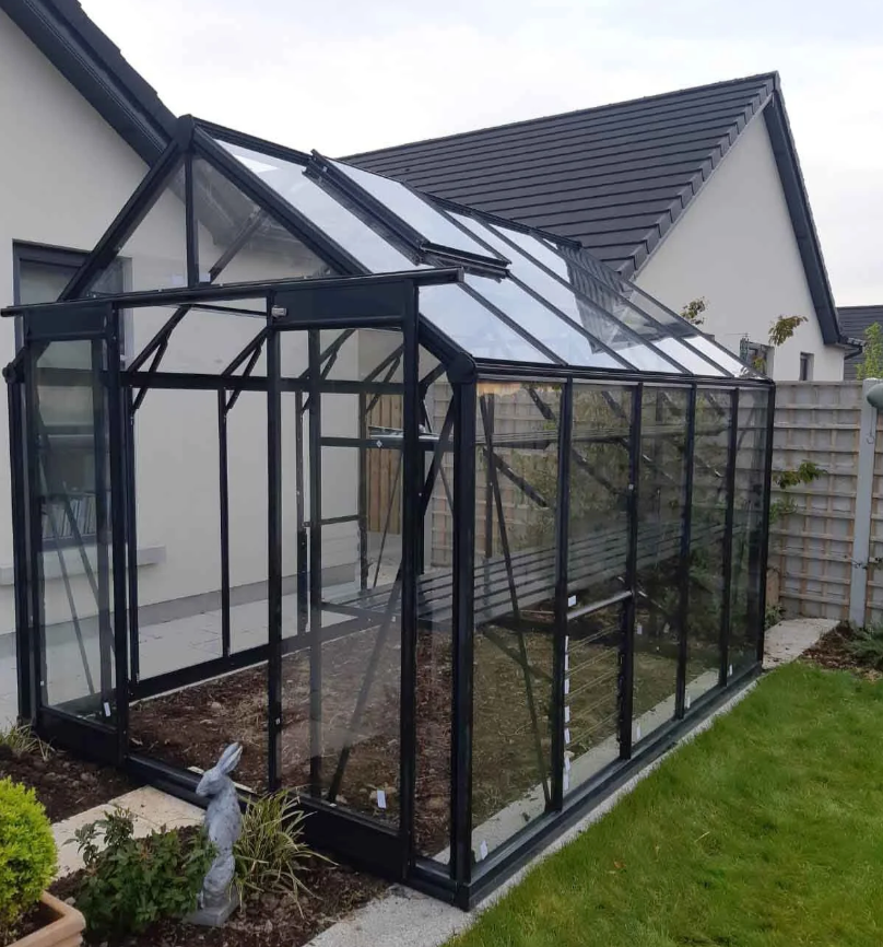 A black-framed glass greenhouse attached to a white house, located in a backyard with grass, plants, and a small garden statue.