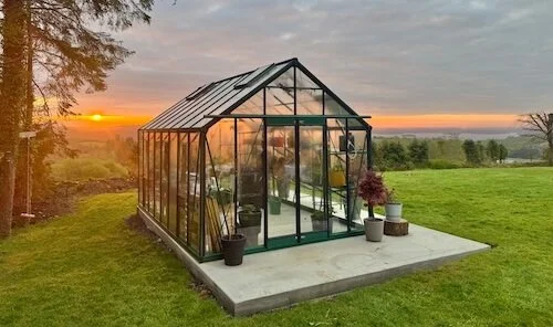 Greenhouse with glass walls and roof on a concrete pad in a grassy yard at sunset, with potted plants outside.